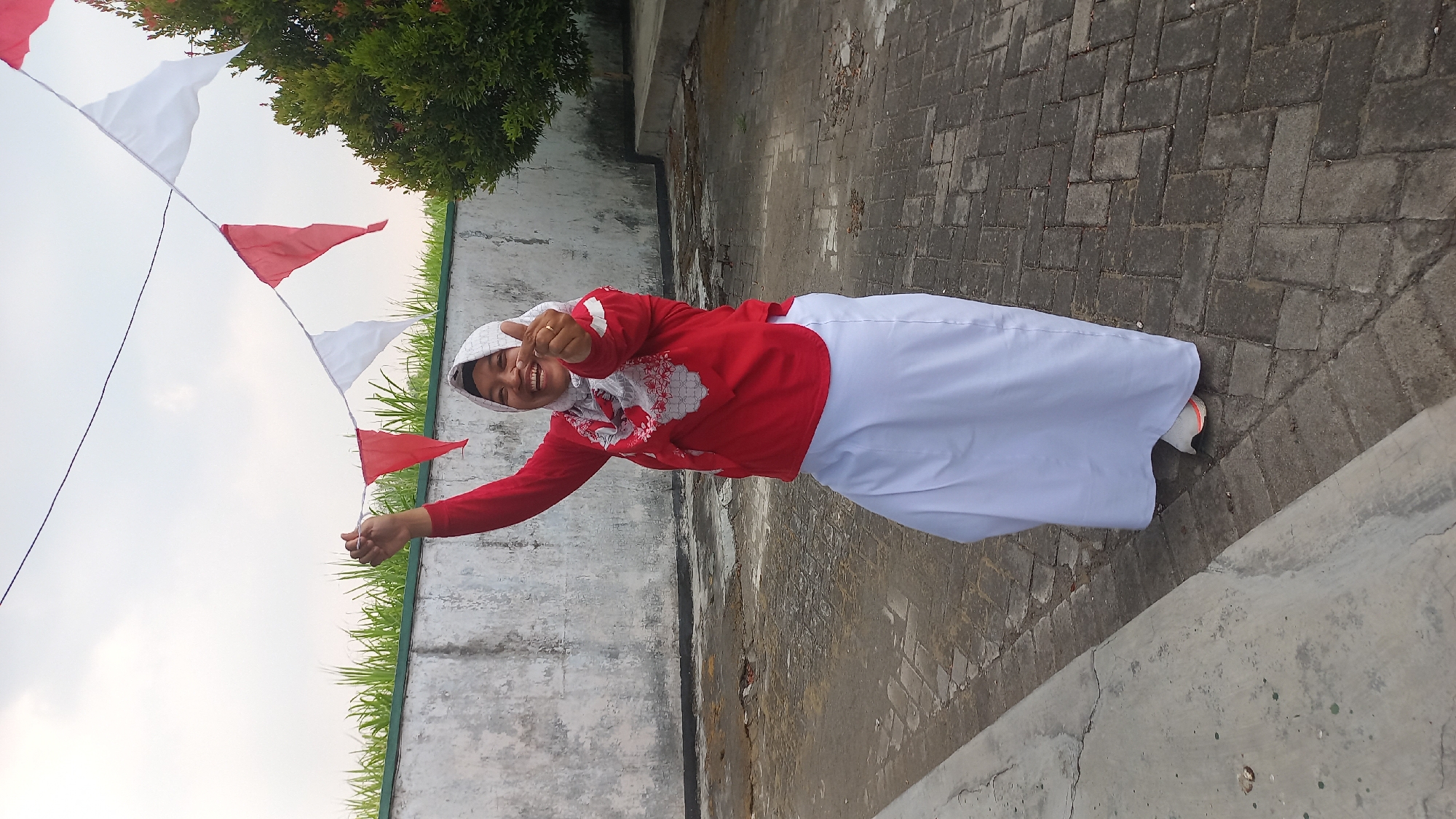 A full-shot photograph depicts a woman, wearing a hijab and a red shirt with white patterned sleeves and a white skirt, standing outdoors in what appears to be a courtyard. She is smiling broadly and giving a thumbs-up gesture with her right hand, while her left arm is raised, holding onto a string of red and white pennant flags. The flags are strung across the frame against a pale, overcast sky. The ground is paved with interlocking bricks. Behind the woman, a low, rough-textured wall is visible, topped by a band of green vegetation. To the right of the woman, a dense green shrub with red flowers is growing. The overall scene suggests a festive or celebratory atmosphere, possibly related to a national holiday given the colors of the flags. The location is identified as Kayen Kidul, Indonesia.