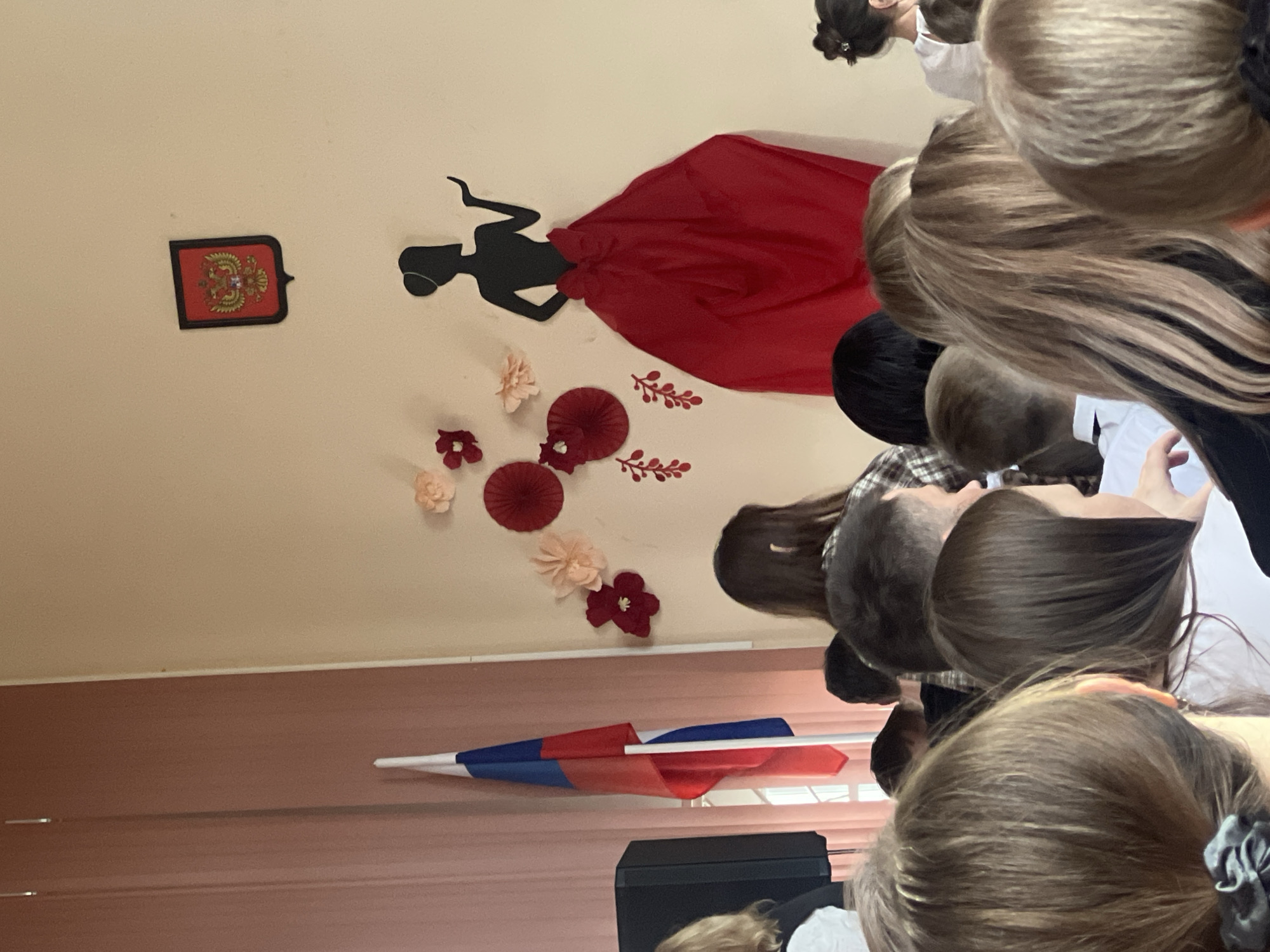 The image, rotated ninety degrees clockwise, captures an indoor scene, likely an event or assembly in a school or community hall in Chaikovsky, Russia. The foreground shows the backs of heads of several young individuals, possibly students, with various hair colors and styles, suggesting they are seated or standing, attentively facing forward.
The light-colored wall is adorned with decorations. A prominent feature is a large black silhouette of a woman in a flowing dress, her skirt dynamically represented by a large piece of vibrant red fabric. Arranged around her are several decorative paper flowers in shades of red, pink, and beige, accompanied by red leaf-like cutouts. To the left of these decorations, a small, framed emblem is mounted on the wall; it features a red background with a golden double-headed eagle design, indicative of a national or regional Russian symbol.
In the lower section of the image, what appears to be the front of the room, a Russian flag with its characteristic white, blue, and red stripes is displayed on a pole, draped against a reddish-brown paneled wall. A black speaker or electronic device rests beneath the flag. The gathered people and the festive wall decorations suggest a school event, a patriotic ceremony, or a cultural celebration. The lighting is even, typical of an indoor setting. No legible text is visible other than the pattern on the emblem.