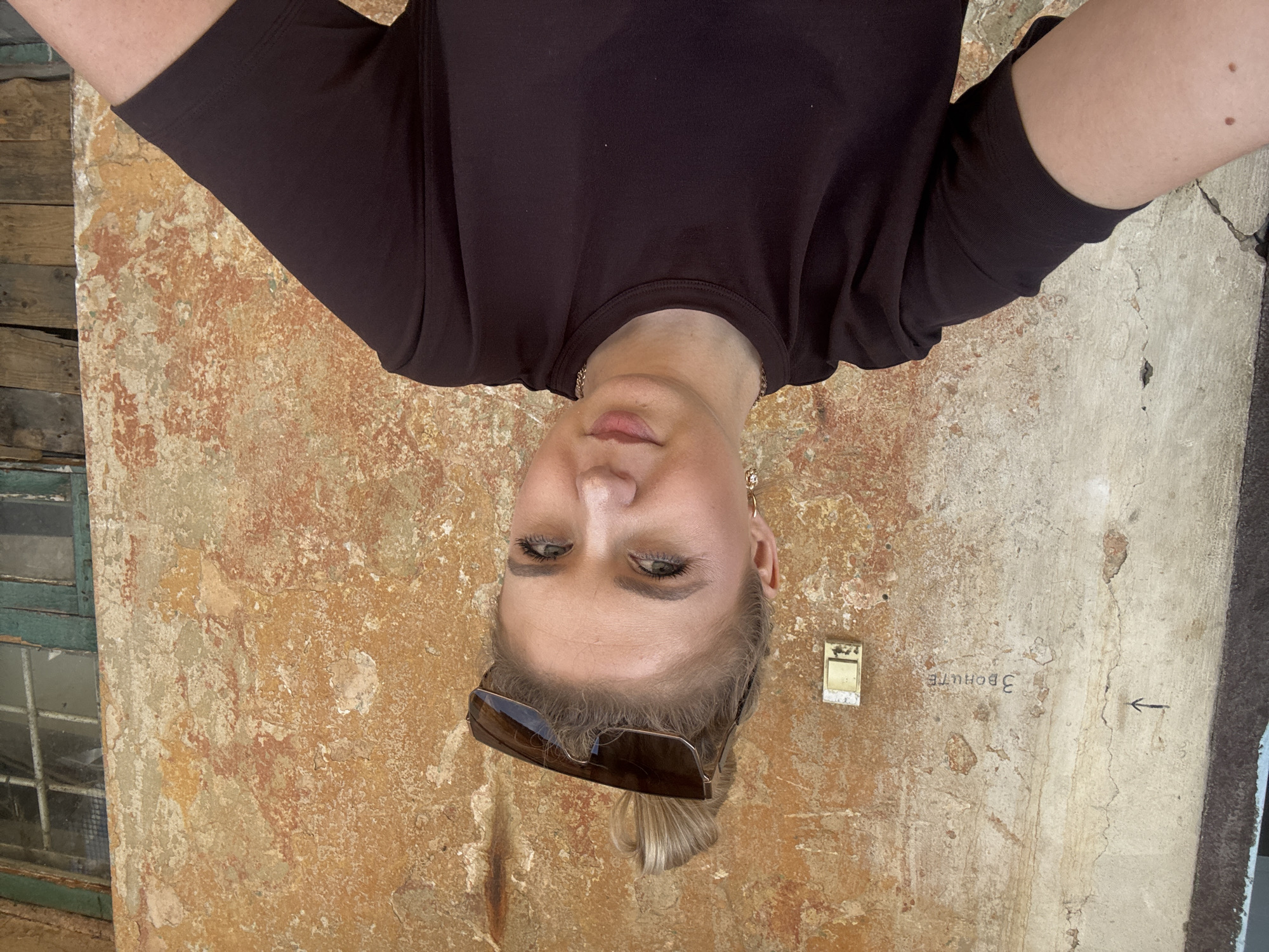 A young woman with blonde hair in a messy bun and sunglasses perched on her head is taking a selfie in front of a textured, peeling wall. She is wearing a dark t-shirt and a delicate necklace. On the wall behind her, a light switch is visible, and to the left, an arrow points towards some faint writing that appears to read "ЗВОНИТЕ" (which translates to "Call"). The wall is a mix of beige, orange, and red hues, with patches of plaster missing, revealing a rougher surface underneath. To the right, a weathered wooden window frame with green paint is visible. The overall environment suggests an outdoor setting, possibly in a historical or aged part of a city.