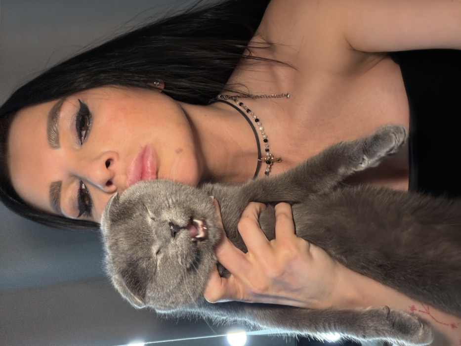 A medium-shot portrait depicts a female individual holding a solid grey Scottish Fold cat indoors, with the scene identified as Svkh Maslovskiy 1 Otdeleniye, Russia. The woman, positioned on the right, possesses long, dark hair and prominent make-up, including winged black eyeliner and filled eyebrows. Her lips are puckered into a 'kissing' gesture, directed towards the cat's head. She wears a black strapless garment and two layered necklaces: an upper black choker with a small golden cross pendant and alternating clear and black beads, and a lower, thinner metallic chain. A small stud earring is visible in her right ear, and a faint red, possibly floral, tattoo is present on her left forearm. Her right hand secures the cat in a vertical orientation. The cat, centrally positioned, exhibits squinted eyes and an open mouth, revealing its tongue and upper and lower teeth in what appears to be a pronounced vocalization or expression. The background is uniformly light grey and out of focus, with several blurred bright light sources visible on the left side.