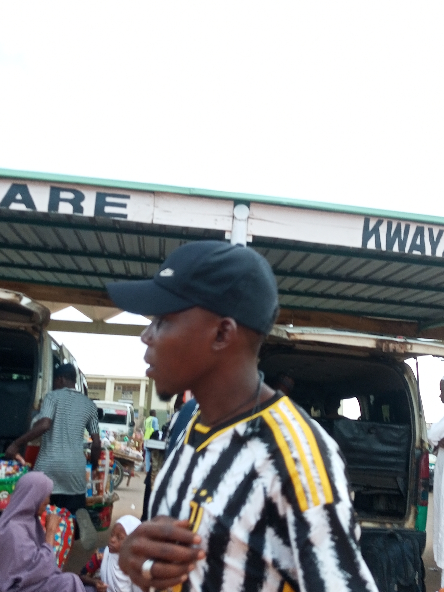 In Gombe, Nigeria, a man wearing a black baseball cap and a black and white striped jersey stands in the foreground. He is facing right, with his mouth slightly open. Behind him, a van is parked with its rear doors open. In the background, people are gathered around stalls and other vehicles, suggesting a busy marketplace or transportation hub. Signs are visible above the vans, one partially reading "KWAYA". The overall scene depicts a public area with commercial activity.