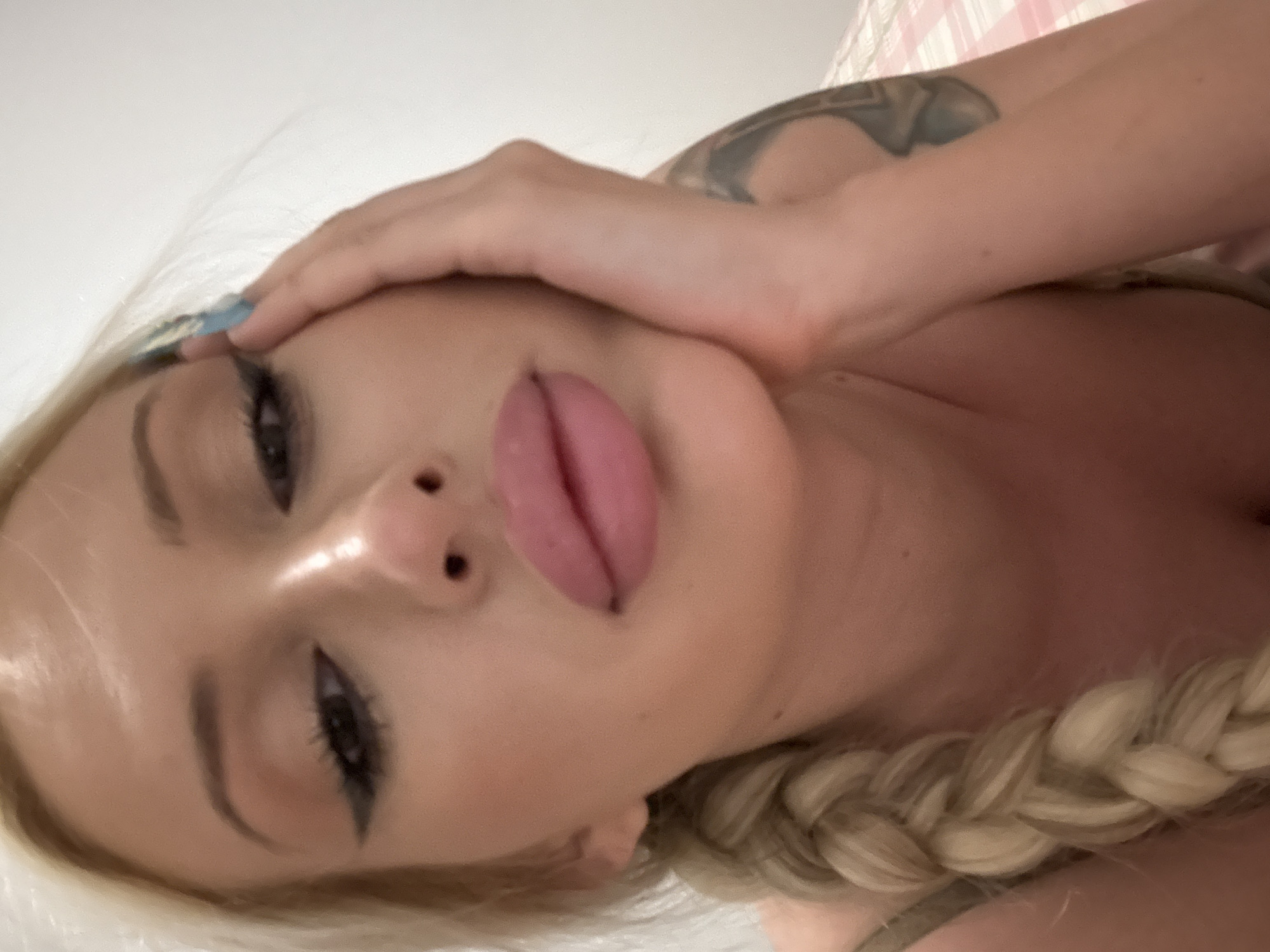 In a close-up, overhead shot, a blonde woman rests her head on her right hand. Her left arm is not visible. She has blonde hair in a braid that rests over her left shoulder. She is wearing makeup, including eyeliner and lipstick. Her right hand is in her hair and partially obscures her face. The background is a light-colored wall.