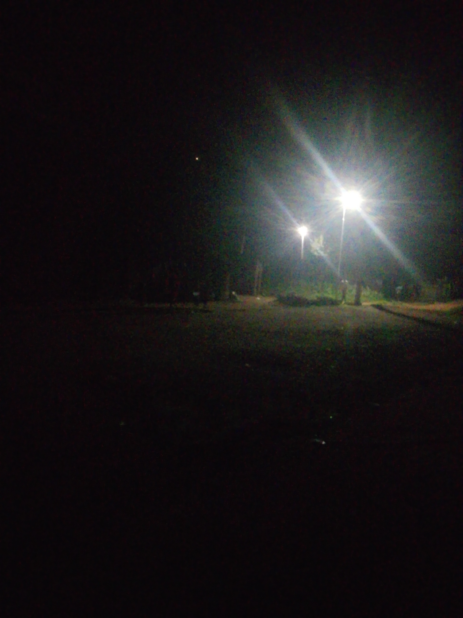 A nighttime scene depicts an outdoor area with two streetlights illuminating a dimly lit path or road. The streetlights are bright and cast beams of light that create lens flares and illuminate the surrounding environment. In the background, trees and vegetation are visible, suggesting a park or a natural setting within or near Kagoma, Nigeria. The ground appears to be covered in gravel or dirt. There are no visible people or animals in the image. The overall impression is one of quiet and solitude, with the artificial light from the streetlights being the primary source of illumination in the darkness.