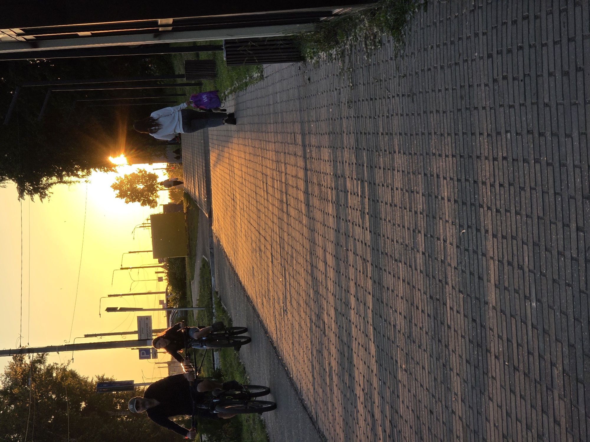 This outdoor image, captured at sunset in Chaikovsky, Russia, depicts a paved pathway bathed in the warm, golden light of the setting sun. The sky is a vibrant yellow and orange near the horizon, with long, distinct shadows stretching across the path from the left.
In the foreground, two individuals are riding bicycles. One male cyclist, wearing a dark top and helmet, looks towards the viewer. Beside him, a female cyclist with long hair, also in a dark top, appears to be looking in the same direction. Further down the path, a person walks away from the camera, wearing a light-colored top and dark pants, carrying a purple backpack with the word "WE" visible on it.
The pathway is constructed from dark, rectangular pavers and is bordered by green grass and trees. In the middle ground, a white street sign with Cyrillic text and a 'P' symbol is visible, indicating "ул. Калинина" (Kalinin Street) and "Приморсkи" (Primorskiy), possibly referencing a "тупик" (dead end). Utility poles and distant urban silhouettes are visible against the bright sky. The scene conveys a peaceful, active evening during golden hour.