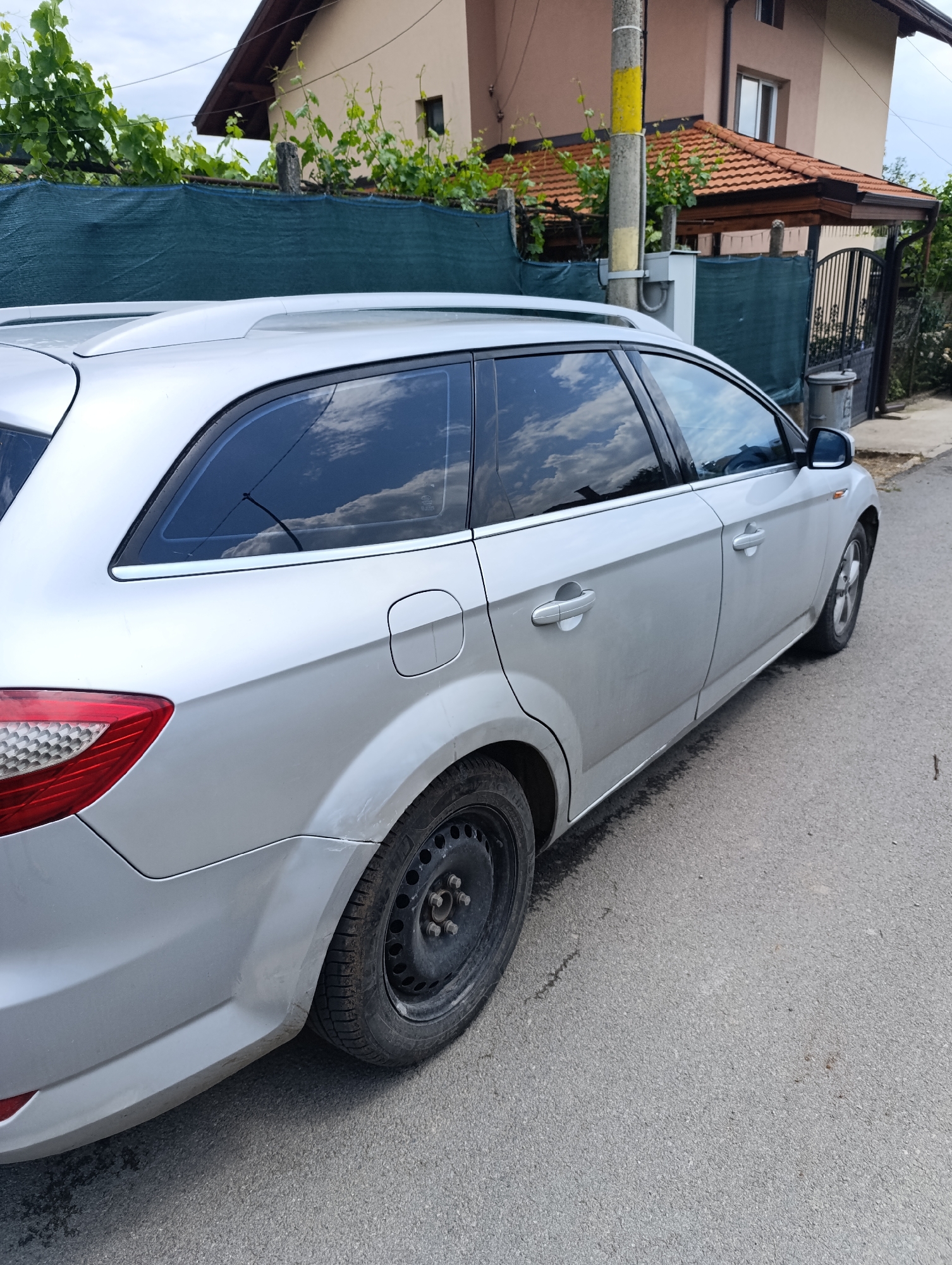The scene depicts a silver Ford Mondeo Mk4 (fourth generation) station wagon parked on an asphalt road in Nikolichevtsi, Bulgaria. The vehicle is positioned with its driver's side facing the camera, angled slightly towards the rear. The car features roof rails and tinted rear passenger and cargo area windows, reflecting an overcast sky with white clouds. The rear driver's side wheel is a black steel rim with a tire, exhibiting dirt and wear. The rear bumper and wheel arch area show minor scuffs and dirt accumulation.
In the background, a dark green mesh fence or shade net runs parallel to the road, partially covered by green vines and foliage. Behind this barrier, a two-story house with a light brown facade and a dark brown tiled roof is visible. To the right of the house, a smaller structure with a red tiled roof extends forward, possibly a carport or covered entrance, adjacent to a black metal gate. A grey concrete utility pole, marked with a yellow band, stands between the car and the house, with utility boxes attached. A grey trash bin is visible near the gate. The sky above is partially cloudy. No people or animals are present in the image.