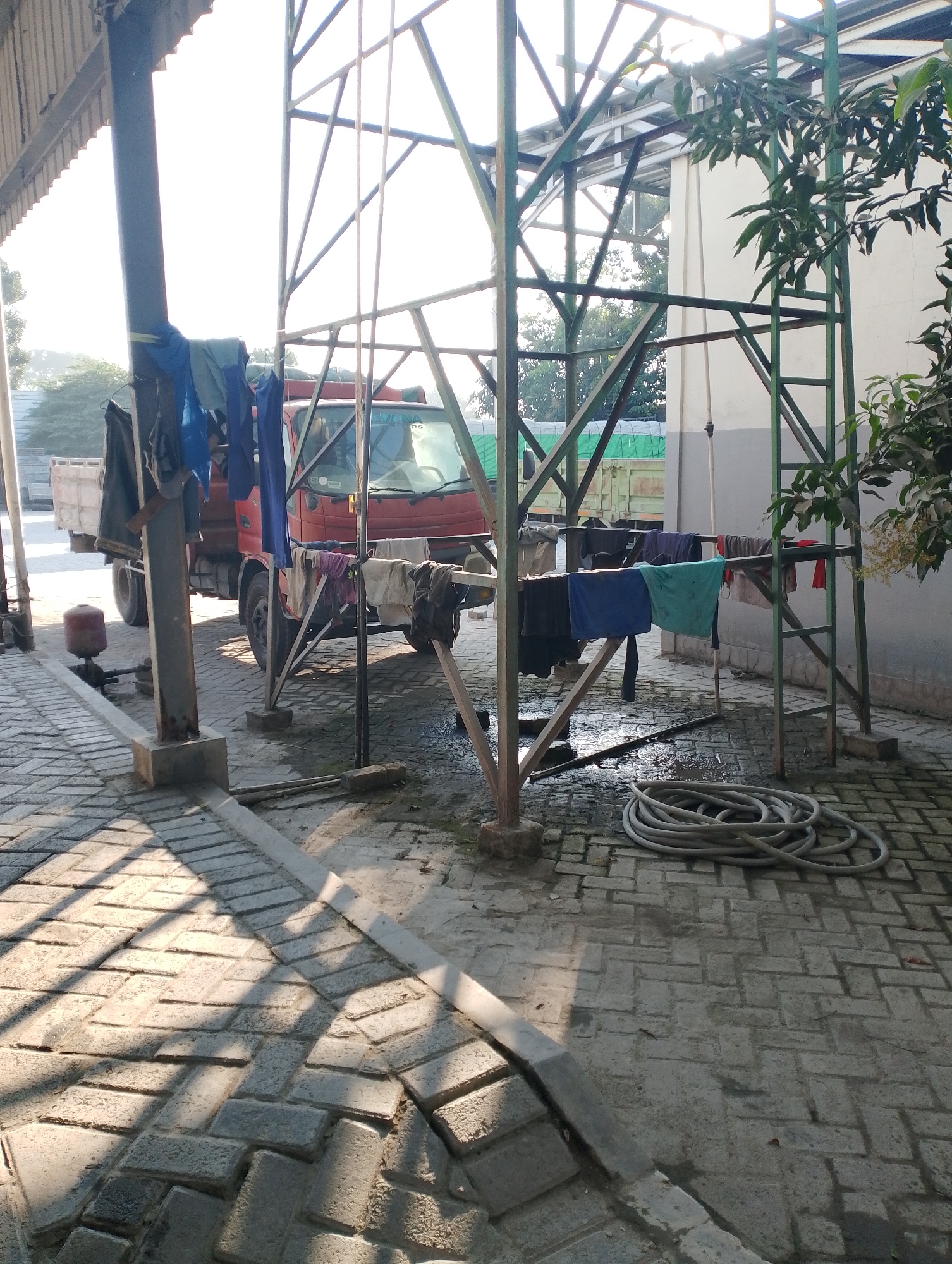 The image shows an outdoor scene in the city of Jambean, Indonesia. A red truck is parked in front of a structure with a green metal frame. Clothes are hanging on a rack in front of the truck. A coiled hose is on the ground to the right of the rack. The ground is paved with interlocking bricks and reflects sunlight, creating a pattern of light and shadow. The scene appears to be in a yard or industrial area. There are no people visible in the image.