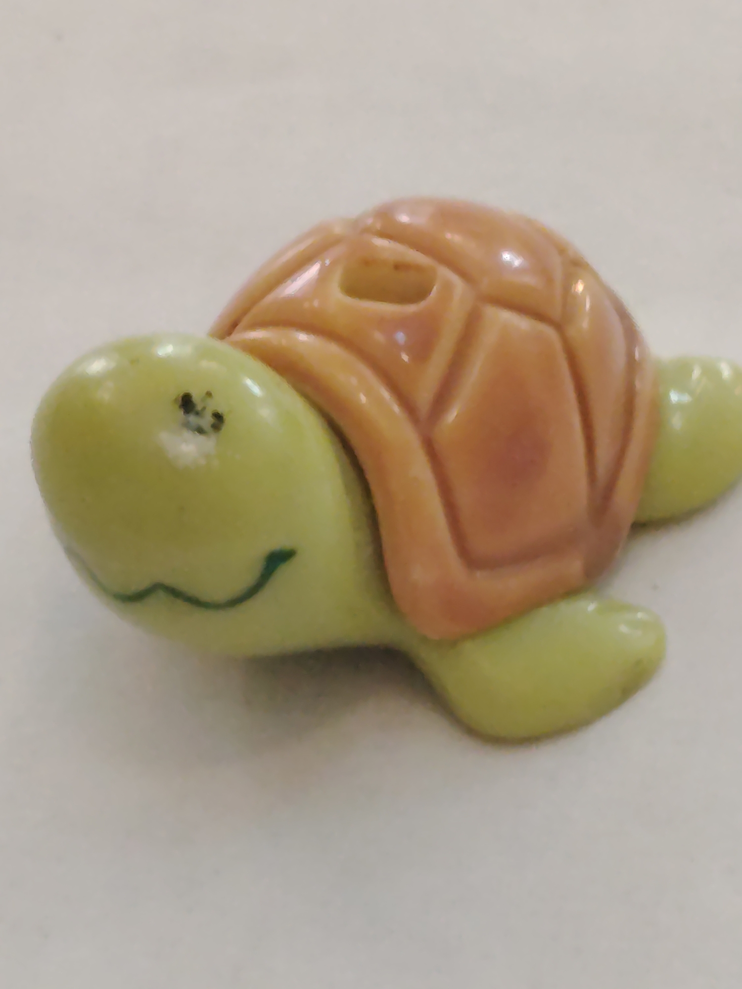 A small, toy turtle rests on a light-colored surface. The turtle has a light green head with a painted green smile and a brown shell with segmented sections. One front flipper and a rear flipper are visible, also in light green. There is no indication of location or any connection to Sarapul, Russia within the image.