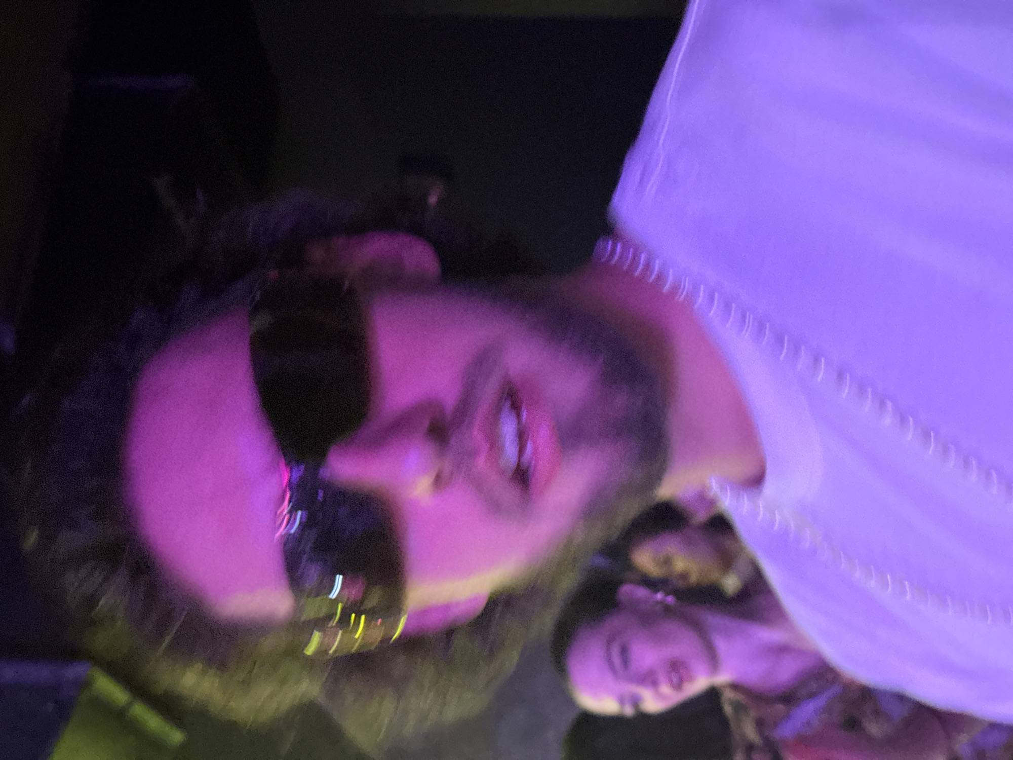 The image shows a close-up of a person with dark hair and a beard wearing sunglasses, bathed in a purple light. Another person is partially visible in the background near the bottom of the frame. A white garment is draped or worn by one of the individuals. The setting appears to be indoors. The city of Sofia, Bulgaria, is not discernable from the visual elements present.