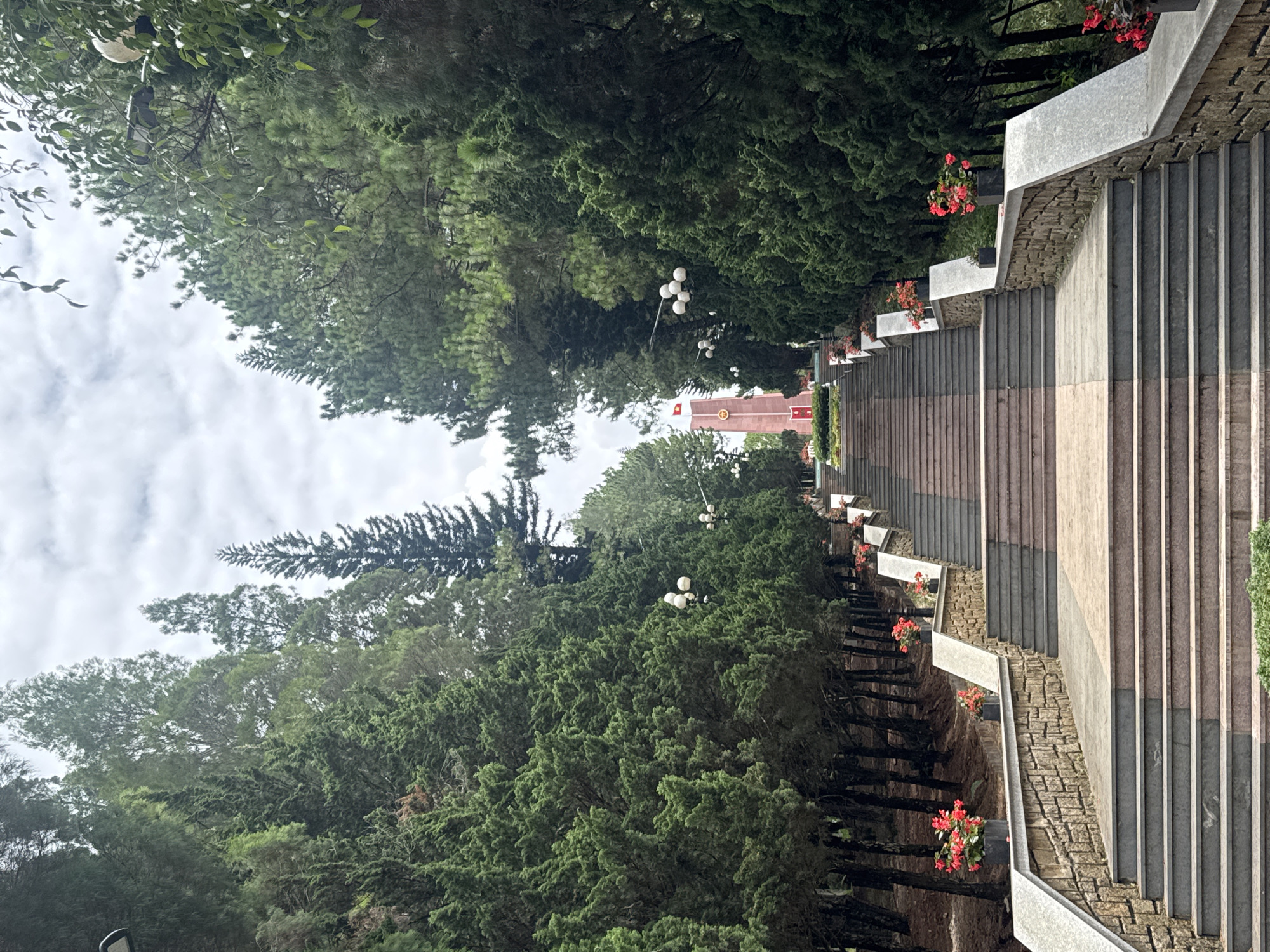 The image shows a wide stone staircase leading up to a pink monument topped with a Vietnamese flag. The staircase is flanked by dense rows of tall evergreen trees. Red flowers in black planters are placed on the retaining walls of the staircase. White globe lights are mounted on poles along the sides of the upper portion of the staircase. The sky is overcast with clouds. The monument appears to be a memorial or historical landmark, consistent with the location of Cam Ly in Da Lat, Vietnam, known for its natural beauty and historical sites. There are no people visible in the image.
