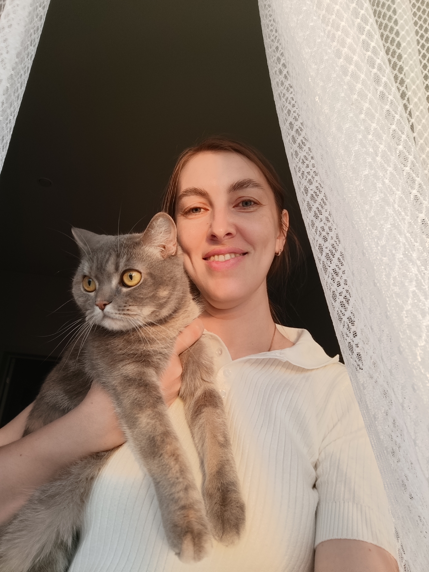 A woman smiles while holding a gray tabby cat. The woman has light skin, brown hair, and is wearing a cream-colored, short-sleeved, collared shirt. The cat has yellow eyes and long whiskers. They are standing in front of a window with a white, lacy curtain. The background is dark, suggesting an indoor location. The scene is bathed in warm light, possibly from the setting sun. There is no information to determine the location in Chaikovsky, Russia.