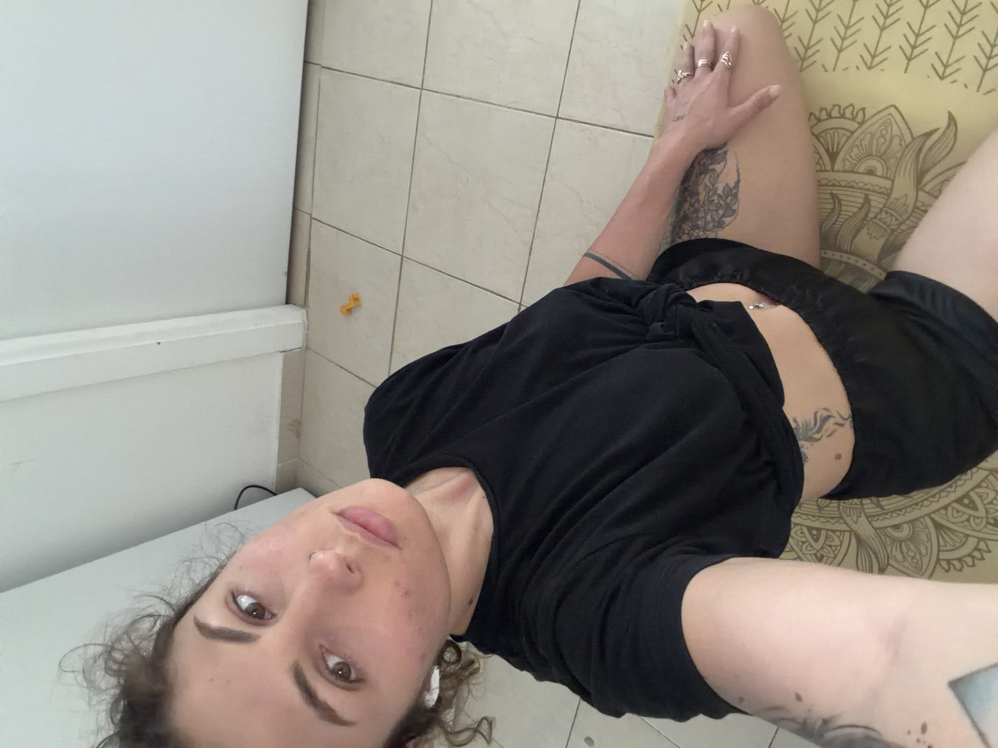 A woman is lying on a yellow patterned yoga mat on a tiled floor. She is wearing a black t-shirt tied at the waist and black shorts. The woman has fair skin, brown eyes, and curly hair. She is wearing rings on her fingers. Tattoos are visible on her upper thighs and arms. A white cabinet is visible along the left edge of the frame. A small yellow object is on the floor near the center of the image. The location is likely a private residence. The city is not discernible from the image alone. Therefore, it is not possible to confirm the location as Kiryat Bialik, Israel.