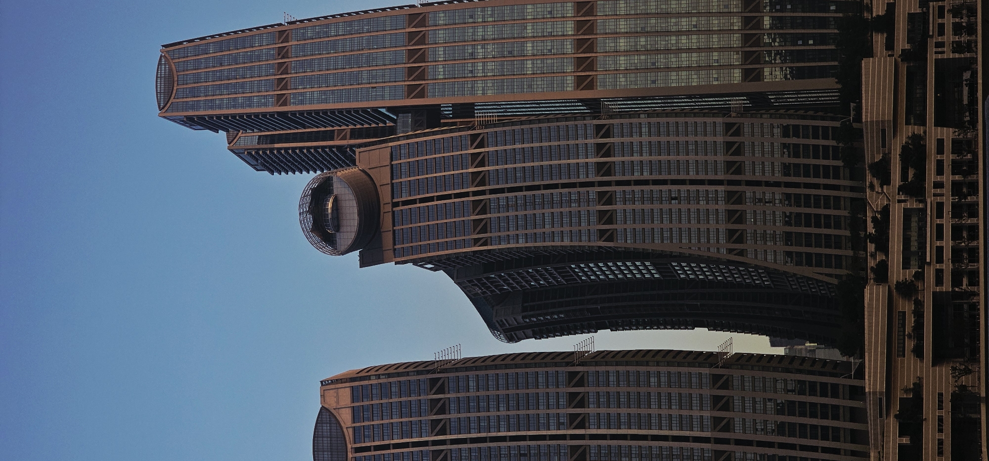 The image shows the distinctive architecture of the Phoenix Islands in Nan'an District, China, against a clear blue sky. The buildings, characterized by their curved, ribbon-like facades and glass exteriors, dominate the frame. The tallest building on the right is a rectangular skyscraper with vertical grid patterns. To its left, a series of interconnected, curving towers create a dynamic, almost fluid silhouette. One of these towers features a prominent circular structure with a glass dome on its upper section, resembling an observation deck or unique architectural feature. A lower, multi-level structure with horizontal banding and greenery adorns the foreground, suggesting a waterfront development or park area. The scene is captured from a distance, likely across a body of water, with the buildings reflecting the soft light of the sky. No people are visible in the image.