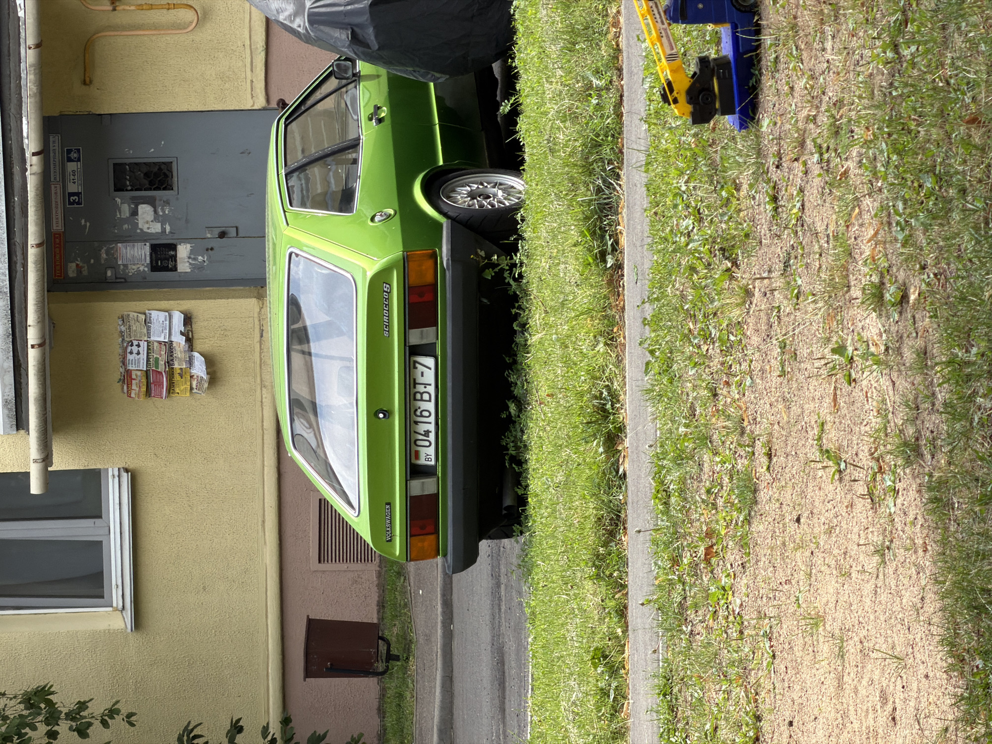 In a residential area of Minsk, Belarus, a bright green vintage Volkswagen Scirocco is parked on the side of the road. The car, with its distinctive sloping rear, is a focal point against a backdrop of a pale yellow building with a grey door and various notices plastered on the wall. A brown trash bin stands to the left of the building. The car is situated on a patch of grass that runs alongside the asphalt road. In the foreground, more grass and a sandy patch are visible, and a bright yellow and blue toy construction vehicle is present on the sandy ground. The scene appears to be a typical street view in a Belarusian city.