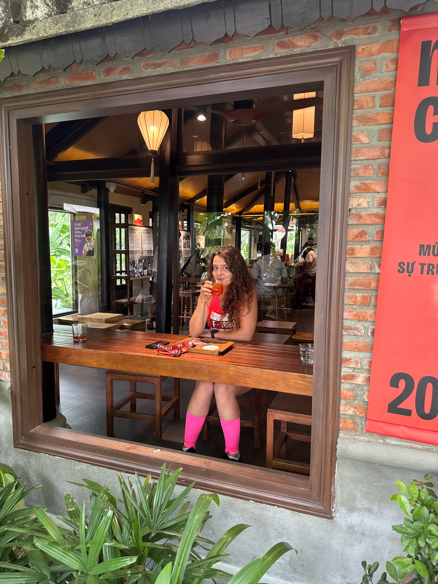 A woman with curly brown hair and a red top is seated at a wooden table outside a cafe in Hoi An, Vietnam. She is holding a drink and looking towards the camera. The cafe has a rustic, traditional Vietnamese aesthetic with exposed brick walls and wooden accents. There are other patrons visible inside the cafe, as well as tropical plants in the foreground. A red banner with Vietnamese text and the number "20" is hanging on the brick wall to the right of the cafe entrance.