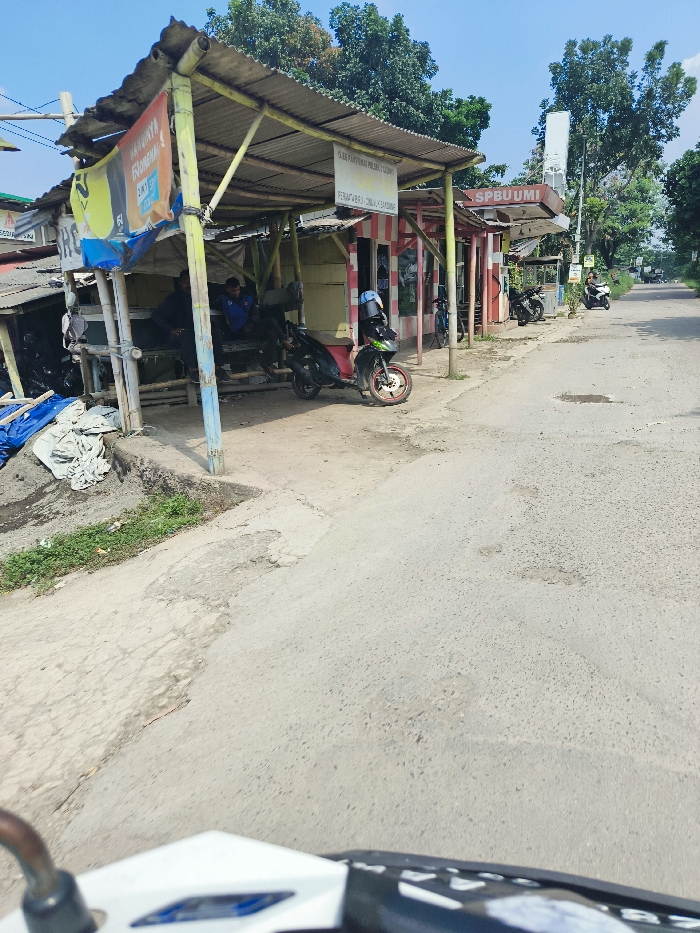The image shows a street in Cileunyi, Indonesia. There are several buildings on the left side of the street with a roof made of corrugated metal, and a few motorbikes parked in front of them. The buildings have a sign that says "SPBU UMI" and "PEMBANGUNAN JALUR PEMBANGUNAN". In the foreground is a white motorcycle, and in the background, the street is lined with trees and a single car is seen in the distance. There are several men in the background sitting under the shelters. The street is made of asphalt and has several potholes. The sky is clear and the weather is sunny.