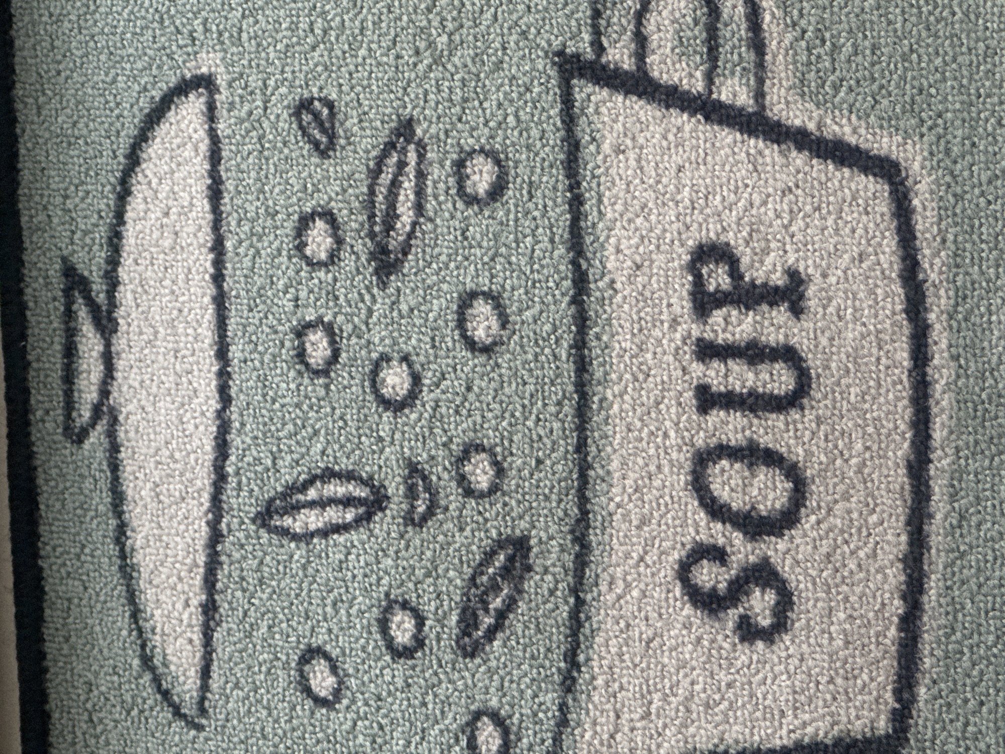 The image shows a section of a rug or mat with a cartoon-style depiction of a pot or container. The pot appears to have a lid and is emitting circular shapes and leaf shapes, suggesting soup or a similar dish. Next to the pot, the word "SOUR" is printed vertically. The background of the rug is a light green color, and the pot and the word "SOUR" are a lighter, off-white color. The setting is not a depiction of an actual location in the city of Puebla, Mexico. It is an abstract image of a cartoon.