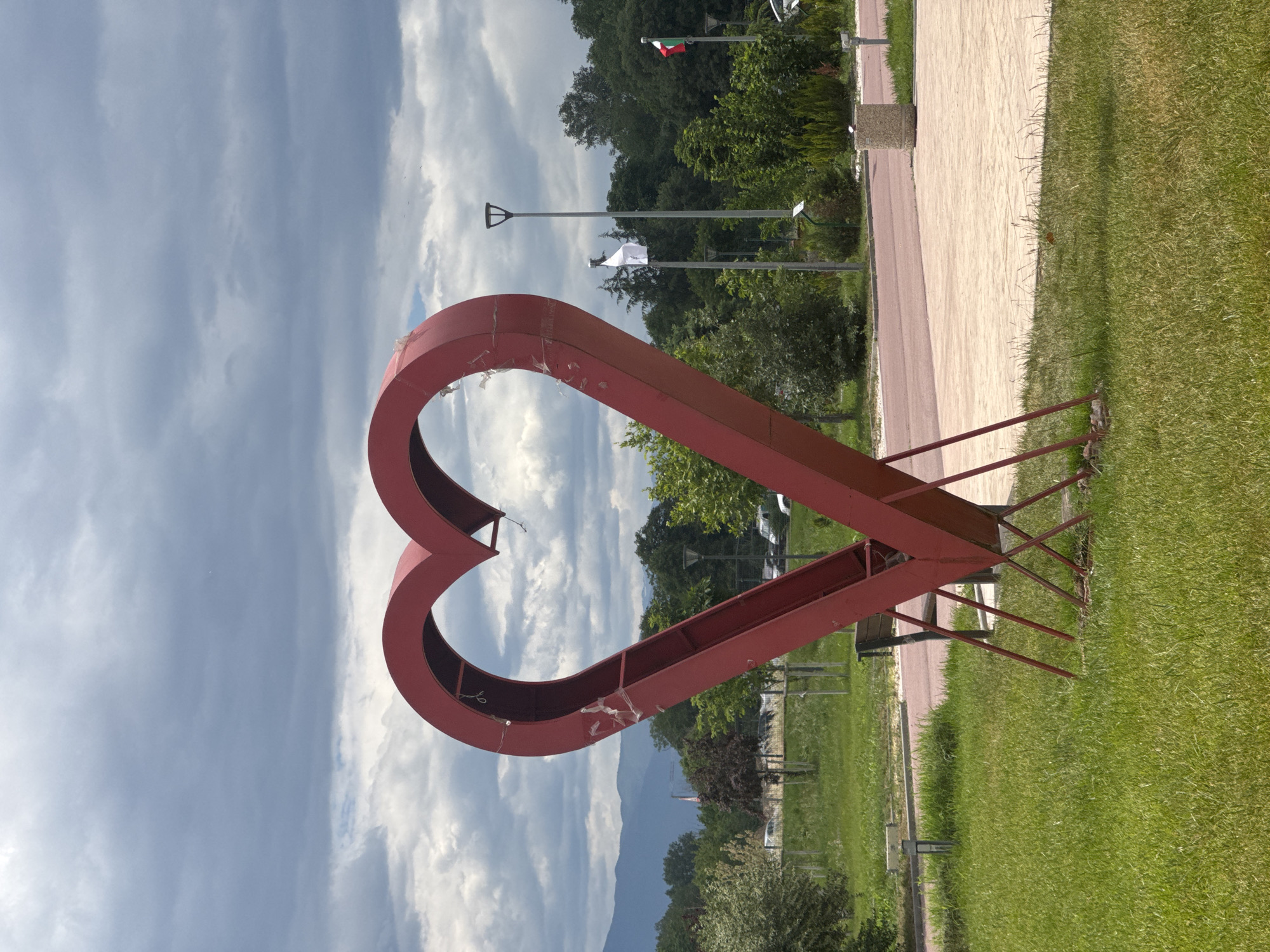 A tilted outdoor shot depicts a large, red metal heart sculpture standing on green grass. The heart is open in the middle, framing a view of trees, mountains, and a cloudy sky. The structure supporting the heart is visible, with metal bars angling into the ground. Adjacent to the grass is a paved walkway. The sky is partially obscured by the heart sculpture, but it shows a mix of blue and white, cloudy formations. In the background, trees line the horizon, and the peaks of mountains are visible. A few streetlights and flags are noticeable in the distance, near the walkway. There is a small strip of greenery between the walkway and the treeline. The photograph appears to have been taken during the day.