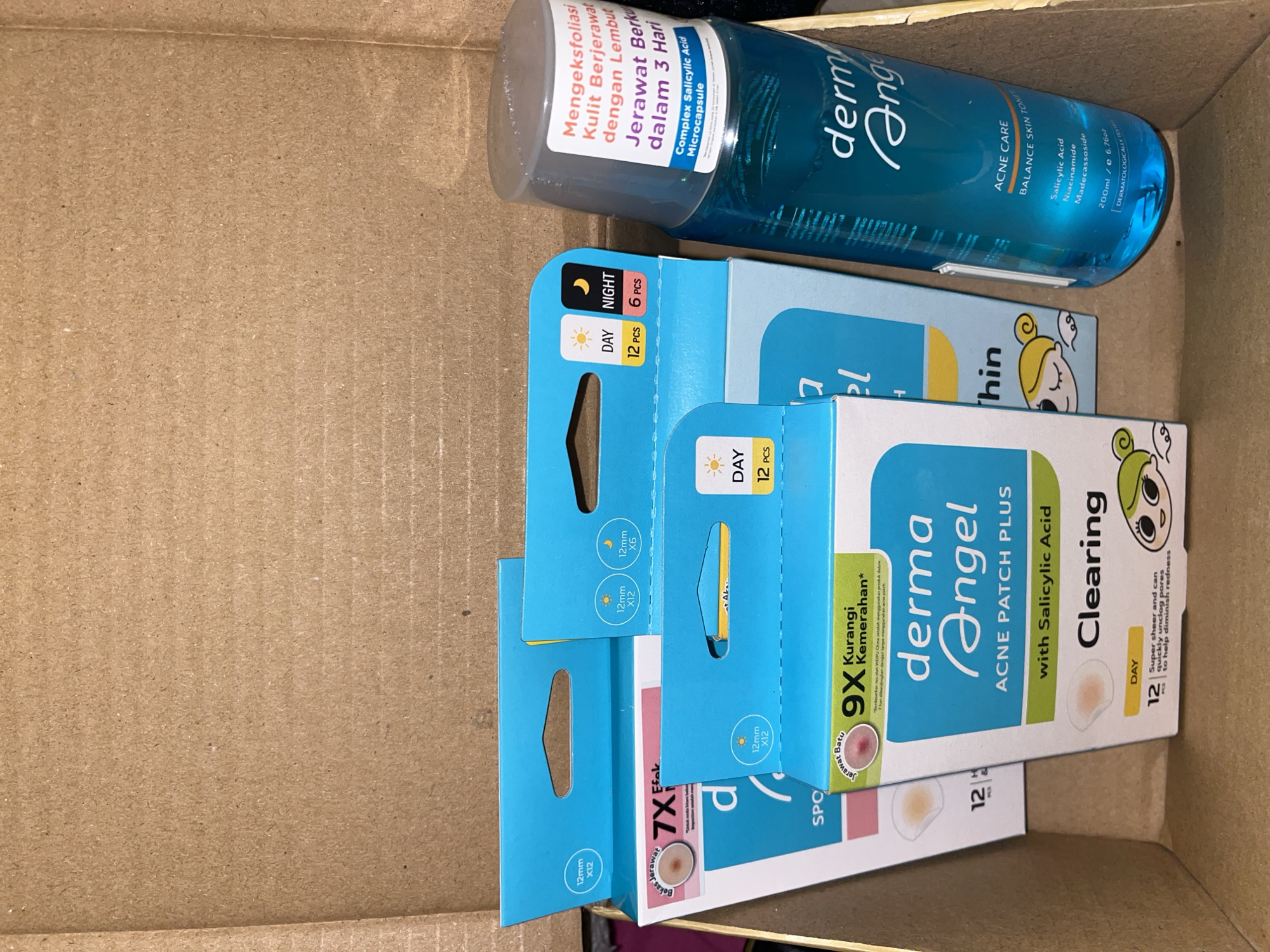 A top-down view shows an open, corrugated cardboard box containing several skincare products. The products are: a blue bottle of derma Angel Acne Care Balance Skin Toner, with visible text in Indonesian referring to exfoliating skin; two boxes of derma Angel Acne Patch Plus with Salicylic Acid labeled "Clearing" and a box of Derma Angel acne patches. The location is unspecified.
