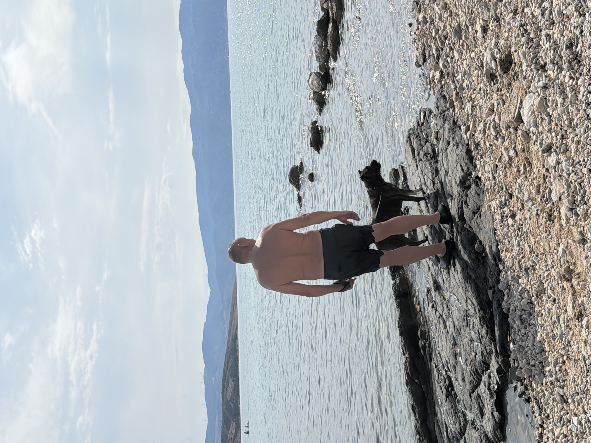A shirtless, adult male stands on a rocky shoreline in Općina Baška, Croatia, facing the bright, calm sea. He is wearing dark shorts and dark slip-on shoes. Beside him, a dark-colored dog, possibly a pit bull breed, stands on the rocks, looking out at the water. The sea is a vibrant blue, with sunlight glinting off its surface, and it extends to a distant, hazy shoreline with rolling hills. The sky above is a pale blue with scattered, wispy white clouds. The foreground consists of various-sized rocks and pebbles, with some smaller stones transitioning to sand closer to the viewer. In the distance, on the water, two people can be seen swimming or wading. The overall scene depicts a peaceful day at the beach, with the man and his dog enjoying the seaside environment.