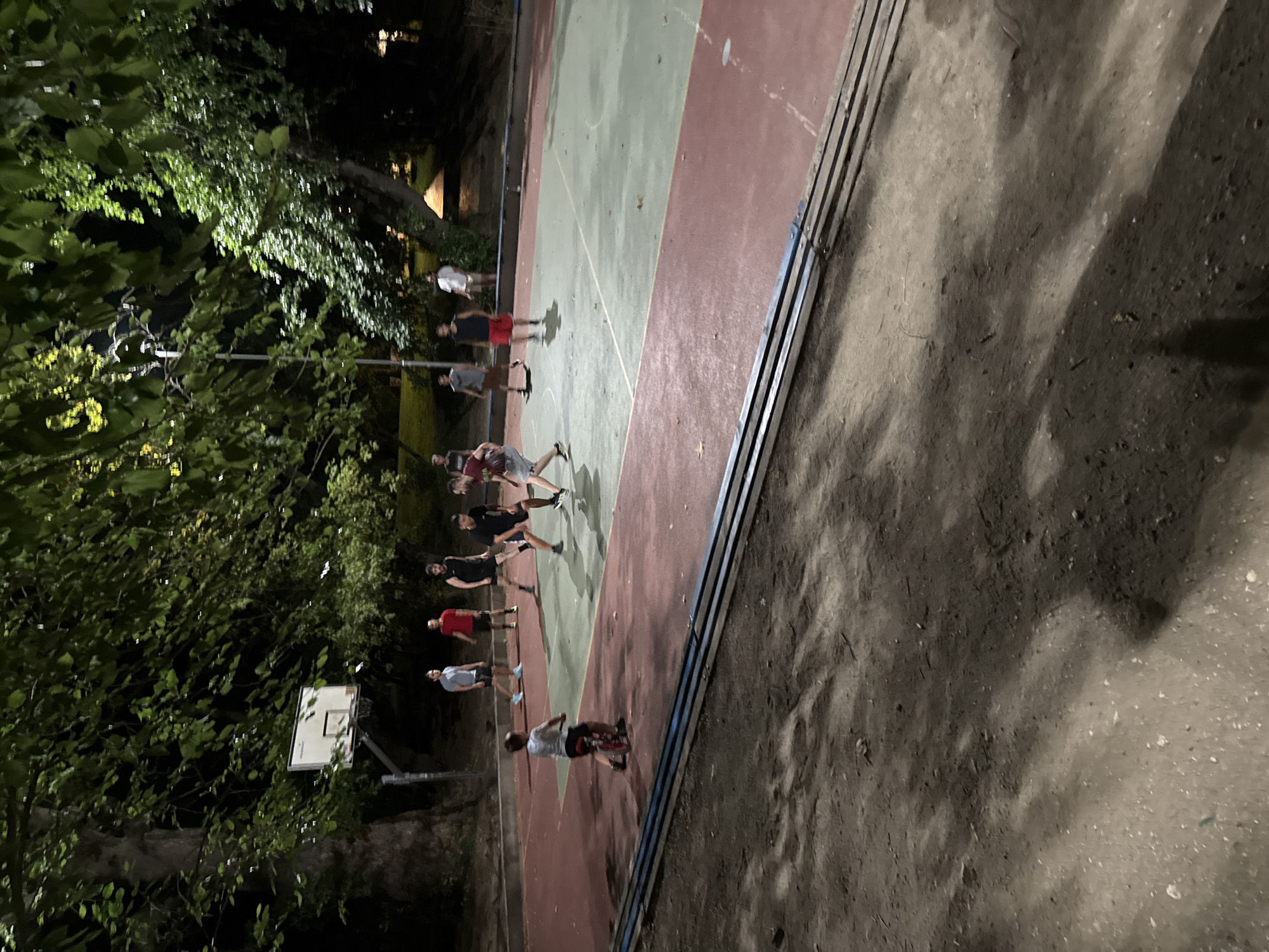 The image depicts an outdoor basketball court at night, illuminated by artificial lighting. Several young men are present on the court, engaged in a game of basketball. One individual is riding a small bicycle on the edge of the court. The court is surrounded by trees, and a basketball hoop with a backboard is visible. The ground surrounding the court appears to be dirt and gravel, with some shadows cast by the trees. The scene suggests a casual, recreational gathering in a park-like setting. This location is identified as the basketball court in the city of Sandanski, Bulgaria.