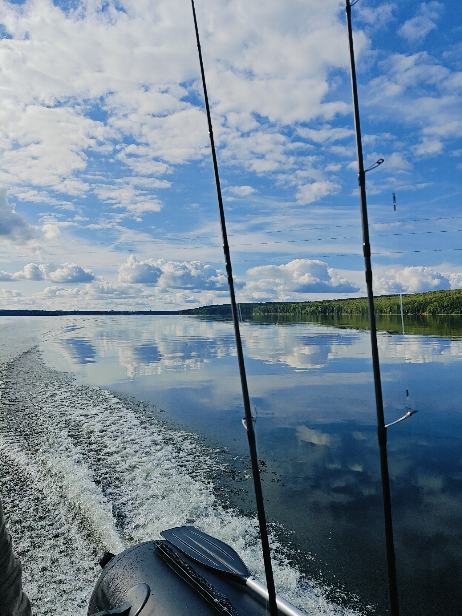 This image depicts a calm body of water, likely a river or lake, with a clear blue sky filled with fluffy white clouds. The water's surface acts as a mirror, reflecting the sky and the treeline in the distance. In the foreground, the wake of a boat is visible, indicating movement. Two fishing rods are prominently positioned in the center of the frame, suggesting a fishing excursion. The scene appears to be set in Chaykovsky, Russia, given the context of the location.