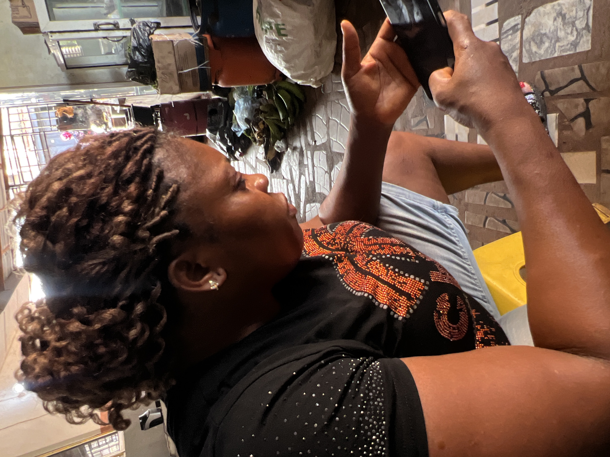 A medium shot captures a Black woman in Nkpor, Nigeria, sitting indoors and looking down at her smartphone, which she holds in her right hand. Her dark, curly hair is pulled back into a bun. She wears a black t-shirt adorned with a pattern of orange and red beads, and light blue shorts. A yellow plastic stool is visible beneath her. The background is cluttered with various items, including stacked boxes, plastic bags, and bunches of green bananas, suggesting a storage or market area. The floor is tiled in a mosaic pattern. Sunlight streams in from the right, casting shadows.