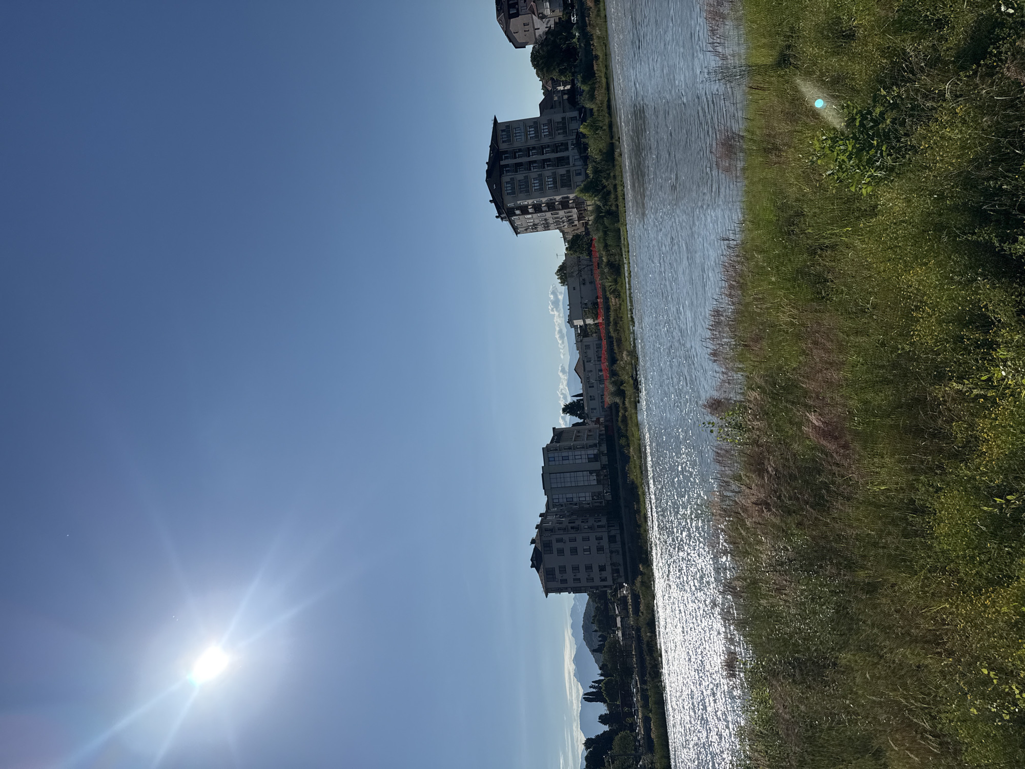 The image depicts a sunlit scene overlooking a wide river, with a city skyline and distant mountains in the background. The sun is bright and high in the clear blue sky, creating a strong glare on the water. The foreground is dominated by lush green grass and vegetation, with some taller reeds catching the sunlight.
On the opposite bank of the river, there are several multi-story buildings, appearing to be residential or office blocks, with a mix of modern and older architecture. Some of these buildings are tall and light-colored, with visible windows and balconies. Further into the background, more urban structures and trees can be seen, leading towards a range of hazy mountains on the horizon. The river itself is calm but reflects the sunlight, creating a shimmering effect.
The scene appears tranquil and urban, with elements of nature in the foreground and distant mountains. The overall impression is one of a pleasant, sunny day in a city situated near a body of water and natural landscapes. There are no people or vehicles clearly visible in the image.