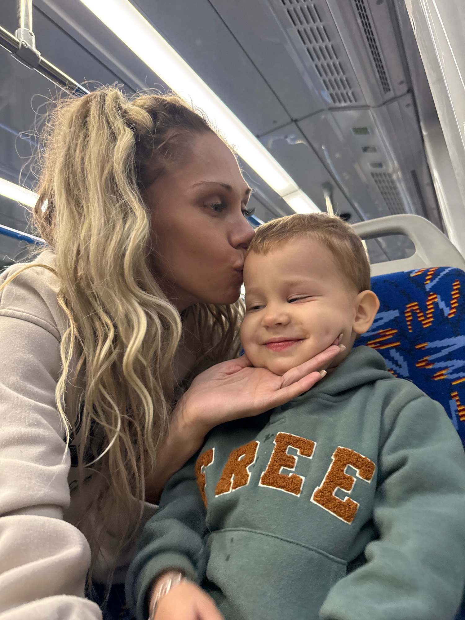 The image captures a tender moment between a woman and a young child, possibly mother and son, on what appears to be public transportation. The woman, with long blonde wavy hair, is leaning in to kiss the child on the forehead. The child, with light brown hair, is looking to his left with closed eyes and a slight smile, his left cheek gently supported by the woman's hand. He is wearing a teal hoodie with the word "FREE" embroidered in orange block letters with a white outline. The woman is wearing a light beige hooded sweatshirt. They are seated on a bench with blue upholstery featuring a pattern of orange and yellow stripes. The background shows the interior of a train or bus, with overhead lighting and ventilation grilles visible. The scene is well-lit, suggesting it was taken during the daytime. The image does not contain any specific visual cues to indicate the location is Yaroslavl, Russia.