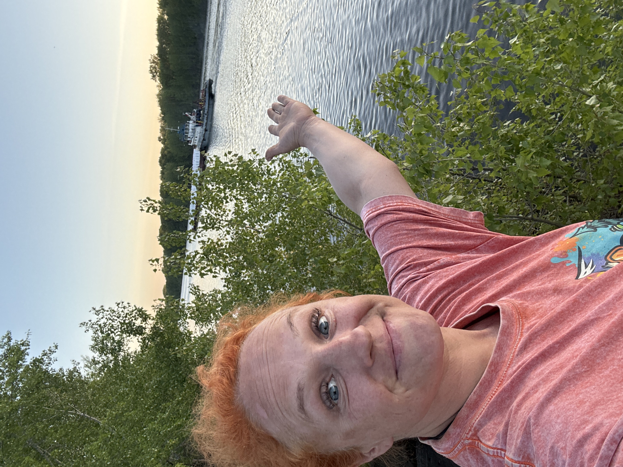 A selfie of a light-skinned woman with red hair taken near a body of water, likely the Kama River, near Chaikovsky, Russia. The woman is wearing a salmon-colored t-shirt. She is holding her right hand up, pointing towards a small boat on the water. There are trees and foliage visible on the bank of the river. The sky indicates it is either sunrise or sunset. The water is rippled.