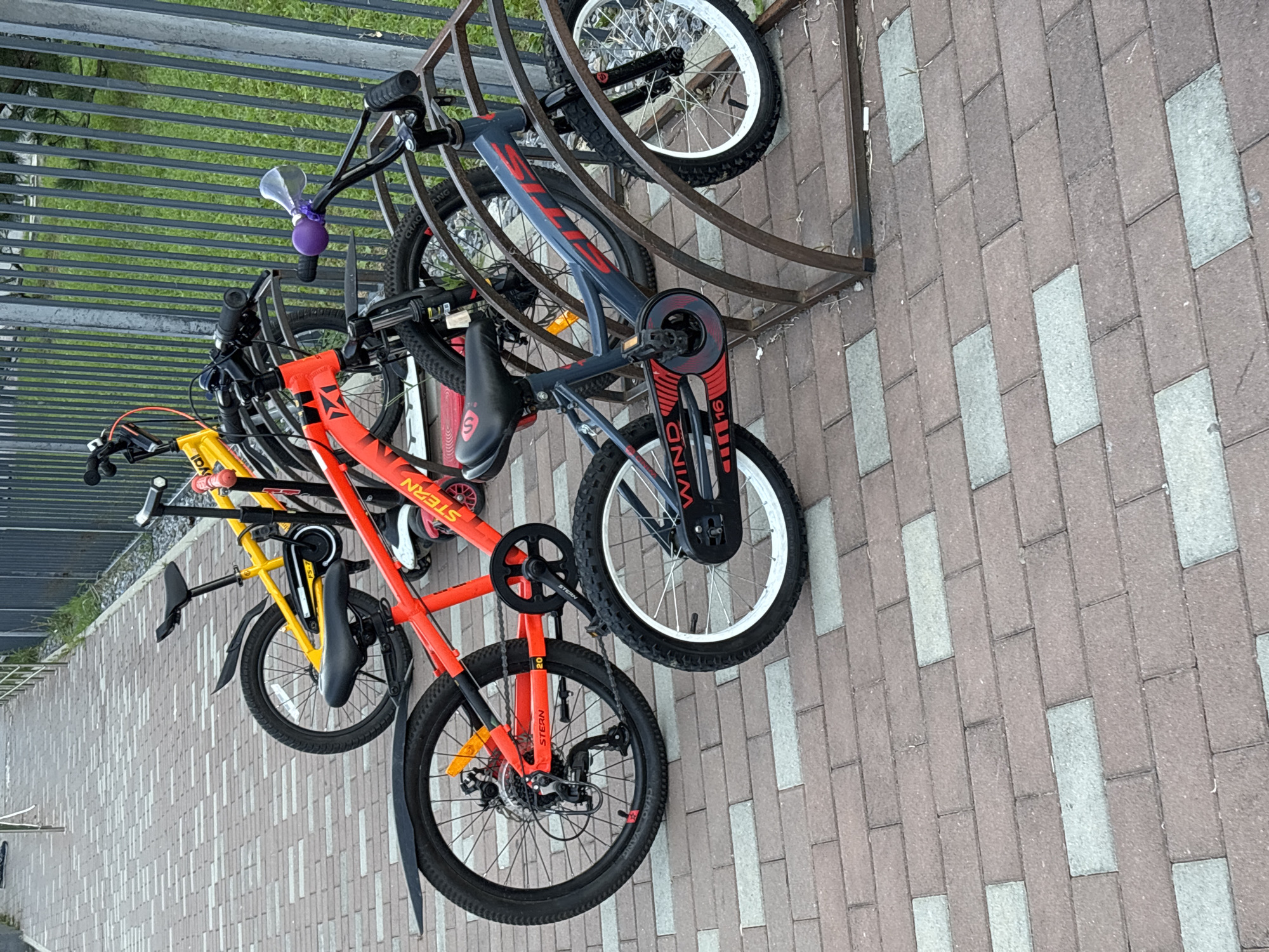 The image depicts a bike rack with several children's bicycles parked in it. The bikes are arranged in a row, with their wheels facing outwards. The bike rack is made of metal and is located on a paved walkway made of brick-like pavers. In the background, there is a metal fence and some greenery. One of the bicycles, a red one, has the brand name "STERN" visible on its frame. Another bicycle, a gray one, has "WIND" written on its chain guard. The scene appears to be outdoors, and there are no people visible in the image. The lighting suggests it is daytime. This scene is a common sight in many urban environments, and without specific visual cues, pinpointing the exact location to Sochi, Russia, is not possible.