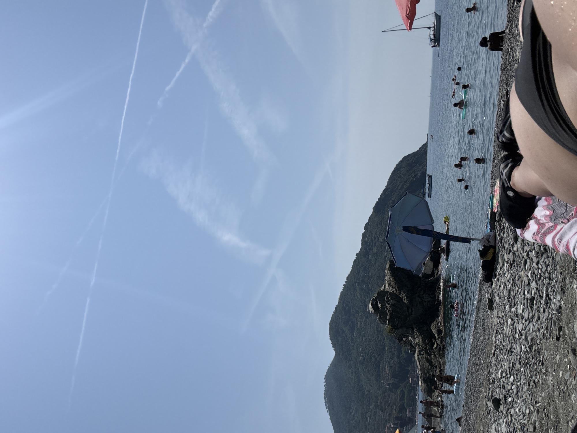 The image depicts a sun-drenched beach scene in Bonassola, Italy, under a clear blue sky with contrails. The foreground is dominated by a person lying on a pebble beach, facing away from the camera, wearing black bikini bottoms. To the left of the person, a striped towel is visible. A blue umbrella is positioned slightly to the left of the frame's center, casting a shadow on the beach and water.

The water is populated with numerous people swimming and floating on inflatable rings and boards. Several figures are visible wading in the shallows. In the middle ground, a large rocky outcrop rises from the sea, with vegetation covering the steep hills of the Italian coastline rising behind it. A white boat is moored near the rocky area.

Further out in the sea, a white sailboat with a red sail is visible on the right side of the image, moving away from the shore. Other distant boats can be seen on the horizon. The overall atmosphere is one of a lively summer day at the beach, with people enjoying the water and the coastal scenery. The presence of contrails in the sky suggests air traffic over the area.