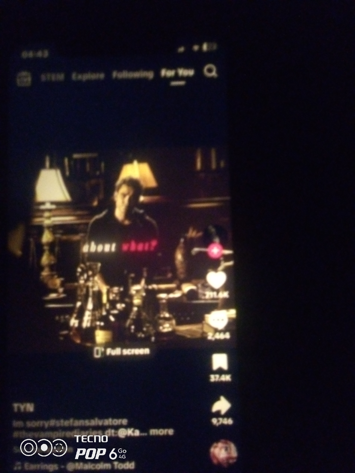 The image shows a close-up of a smartphone screen displaying a video. The video features a person wearing a black t-shirt with the words "about what?" printed in white and red. The background of the video appears to be a dimly lit room with lamps and furniture. On the right side of the screen, there are social media interaction icons such as a heart and a speech bubble, along with corresponding numerical counts. At the bottom of the screen, there is text that includes hashtags and the name "TECNO POP 6 Go 4G", suggesting the video is from a platform like TikTok. The image quality is poor, with blurriness and low resolution, making it difficult to discern finer details. The scene does not provide any specific visual cues to determine the location in the city of Howayi, Nigeria.
