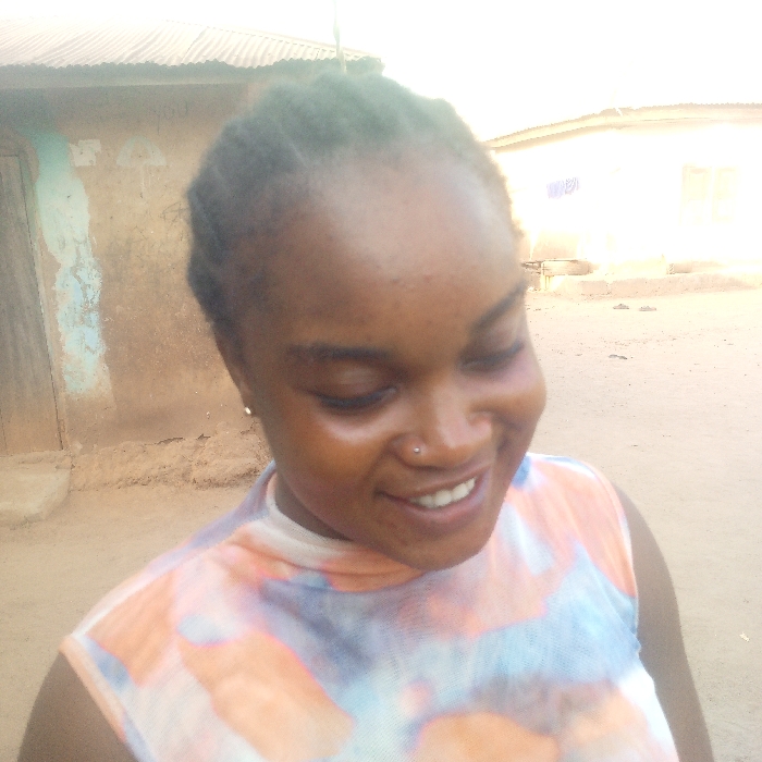 This image captures an outdoor scene in Igbreda, Nigeria, featuring a young adult female in a close-up, slightly off-center to the left. The female subject has dark skin, dark braided hair, and a small silver stud nose piercing on her left nostril. Her eyes are closed or cast downwards, and she is smiling, revealing her upper teeth. A small silver earring is visible in her right ear, and her skin exhibits a natural sheen on her forehead and cheeks. She wears a sleeveless, high-neck top patterned with white, light blue, and orange hues.
The background on the left consists of a building with a weathered corrugated metal roof, a rough, earth-toned wall, and a visible dark brown door. A faded light blue marking is present on this wall. Further to the right in the background, a lighter-colored building with a window is partially visible. The ground appears to be dry, dusty earth. The scene is illuminated by natural daylight.