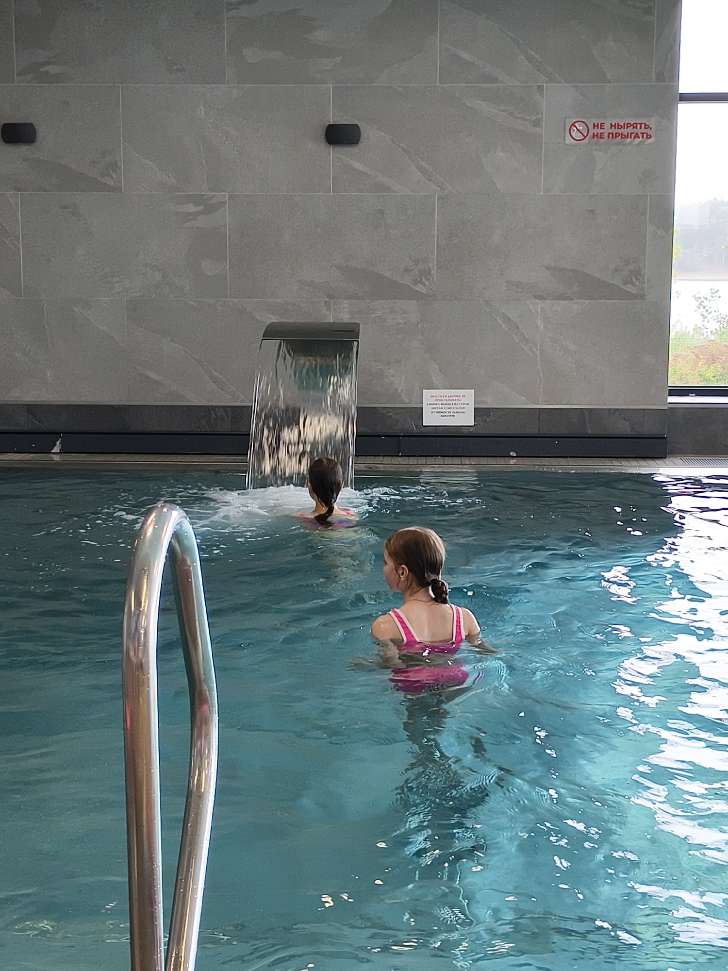 In the city of Chaikovsky, Russia, two young girls are swimming in a modern indoor pool. One girl, with dark hair in a ponytail, wears a pink tankini and is in the foreground, facing away from the camera. The other girl, also with dark hair, is further back in the pool, near a waterfall feature, and is also facing away. The pool has clear blue water and is surrounded by grey tiled walls. A metal railing is visible in the bottom left of the frame. A small sign on the wall above the pool has red text with a prohibition symbol, indicating rules for swimmers.