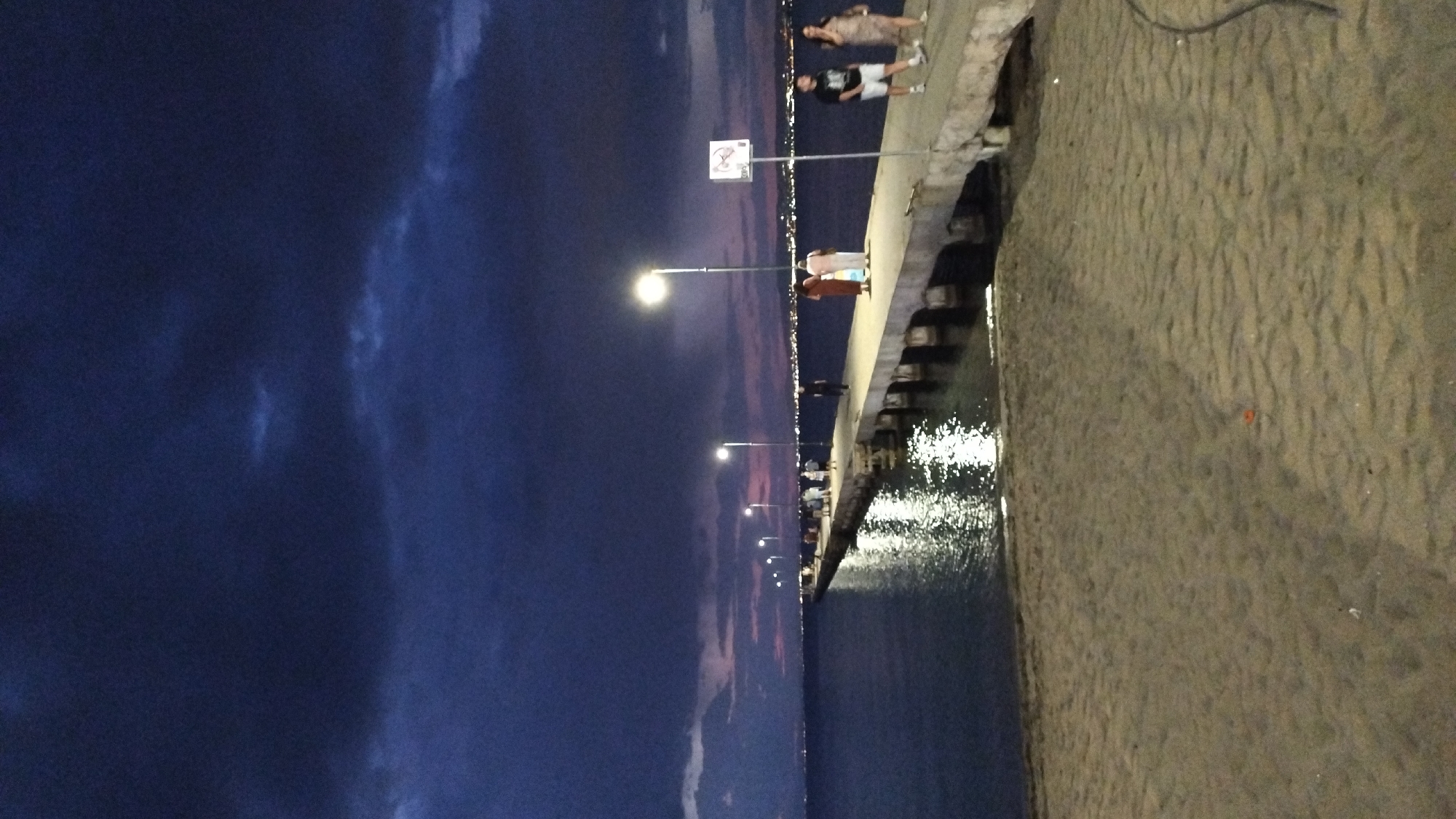 A long concrete pier extends into a body of water from a sandy beach at dusk. The sky is a deep blue with dark, heavy clouds, and a faint pink and orange hue is visible on the horizon, suggesting the last light of sunset. Several streetlights line the pier, casting pools of light onto its surface and reflecting brightly on the water below. A few people are visible on the pier, walking or standing. On the far right, two individuals are walking towards the foreground, and further down the pier, a small group of people are gathered. In the distance, the lights of a city twinkle across the water. The beach in the foreground is composed of light-colored sand with some rippled texture. The water appears dark and calm.