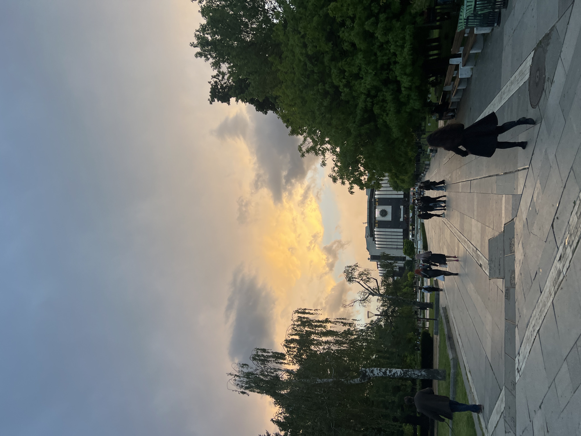 The image shows a street scene in Sofia, Bulgaria. The sky is a mix of gray and golden clouds, indicating sunset or sunrise. In the middle distance, people are walking along a paved walkway toward a modern building with dark windows. The building has a geometric design and appears to be a public structure. On the left and right edges are trees, some birch and some larger, denser foliage. On the right edge of the walkway, benches line the path. One person in a dark coat and dark hair is walking away from the viewer on the right side of the path. Another person is walking toward the camera on the path's left side. The scene suggests an urban environment during dusk or dawn with people strolling leisurely. Given the architectural style and surroundings, the location is likely near the National Palace of Culture (NDK) in Sofia.