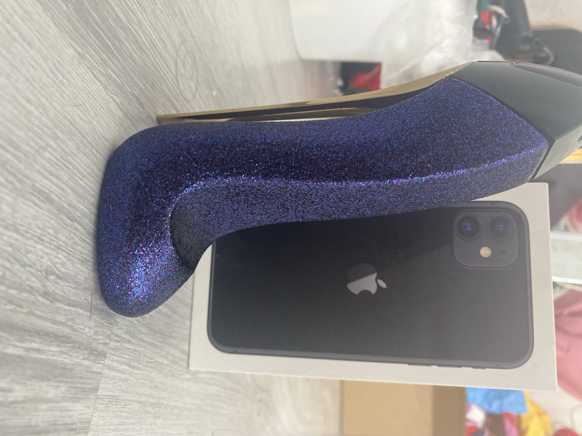 A close-up, low-angle shot shows a sparkly purple stiletto heel next to a black iPhone 11 box. The stiletto is positioned in the foreground, angled towards the right, with its pointed toe facing the viewer. It has a high, slender gold heel. Behind the stiletto, a white box with the Apple logo and the outline of an iPhone is visible. The background is blurred, but appears to be an indoor setting with hints of other objects and possibly shelves. The floor is a light gray wood-grain laminate. The overall lighting is soft. The image does not contain any contextual information about its location in Blagoevgrad, Bulgaria.