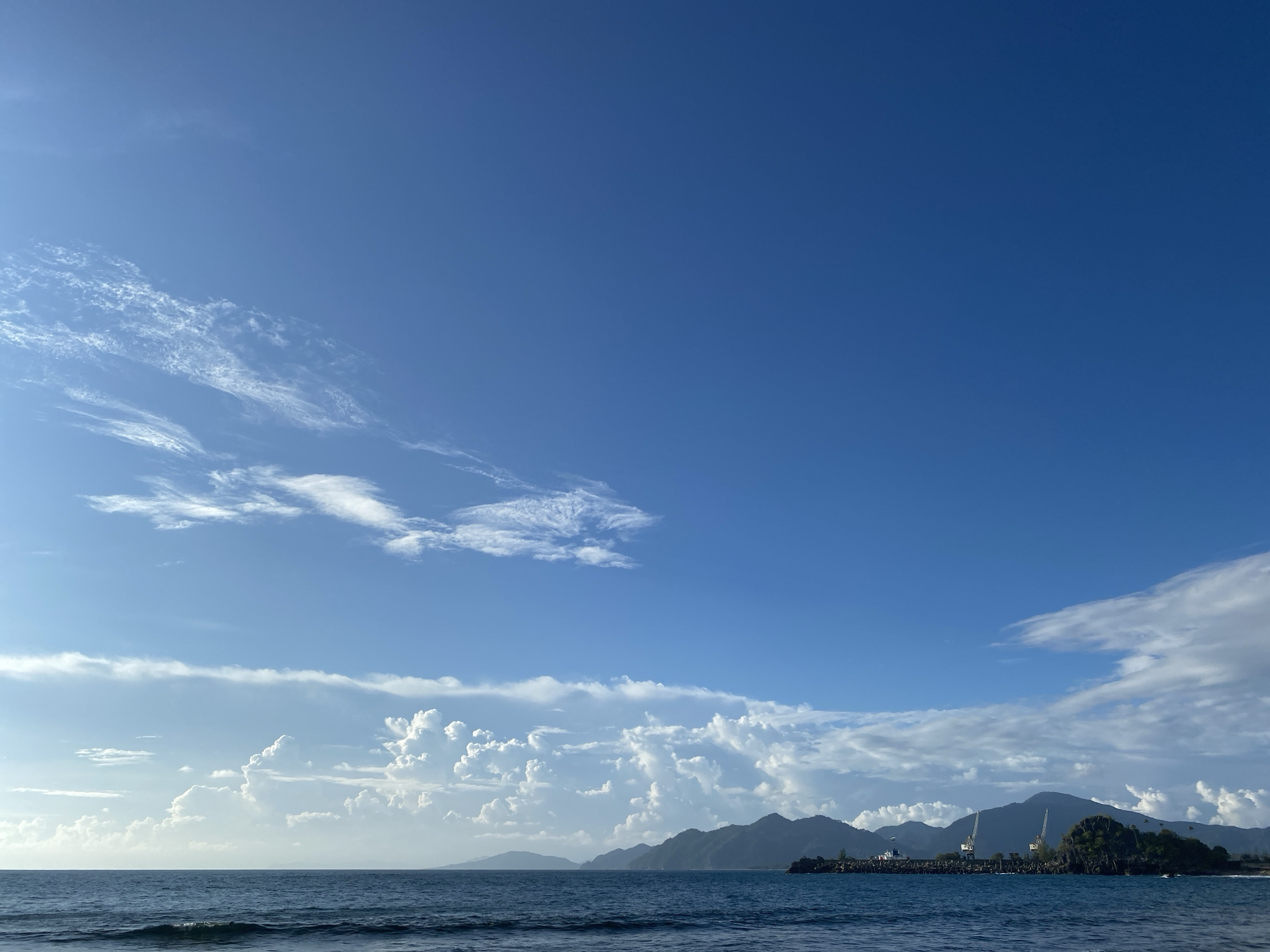 The image depicts a coastal landscape under a vast blue sky with scattered white clouds. In the foreground, the deep blue ocean stretches across the frame with gentle waves. In the mid-ground, a rugged coastline is visible, dotted with mountains in the distance. A prominent man-made structure, appearing to be a pier or breakwater, extends into the water, featuring several cranes. The scene is set in Monikeun, Indonesia.