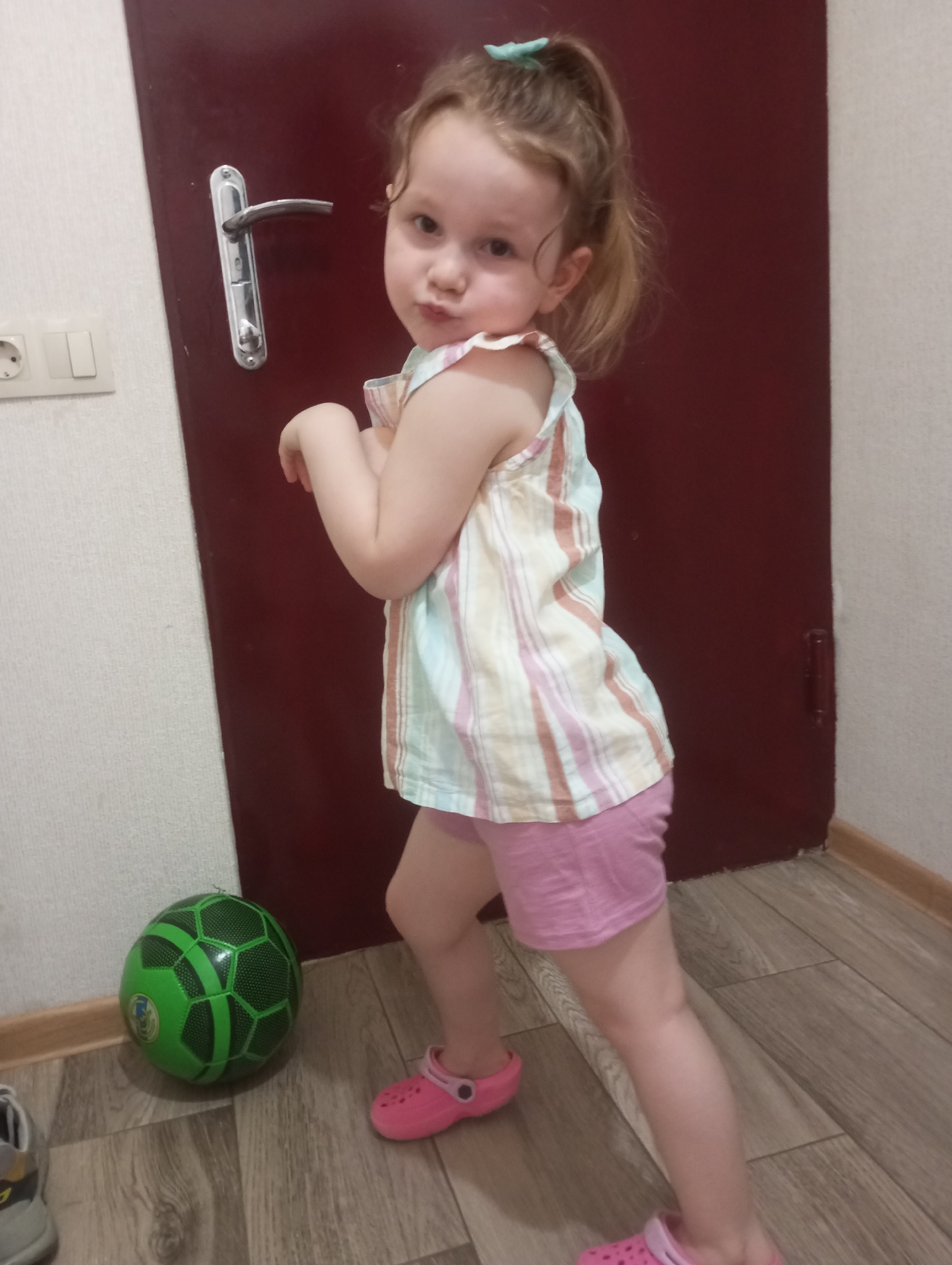 A young Caucasian girl with blonde hair pulled into a ponytail and a light blue hair clip stands on a wooden floor. She is wearing a striped tank top with pastel pink, yellow, and light blue stripes, and light pink shorts. Her arms are crossed in front of her chest. To her left, a green soccer ball with black hexagonal patterns rests on the floor. To the far left, a gray shoe with yellow accents is visible. Behind the girl is a dark red door with a silver handle, and to the left of the door is a white wall with an electrical outlet and light switch. The overall scene appears to be indoors, possibly in a hallway or entryway, and it is unlikely to be a specific location within Tbilisi, Georgia, as no defining landmarks or identifying features are present.
