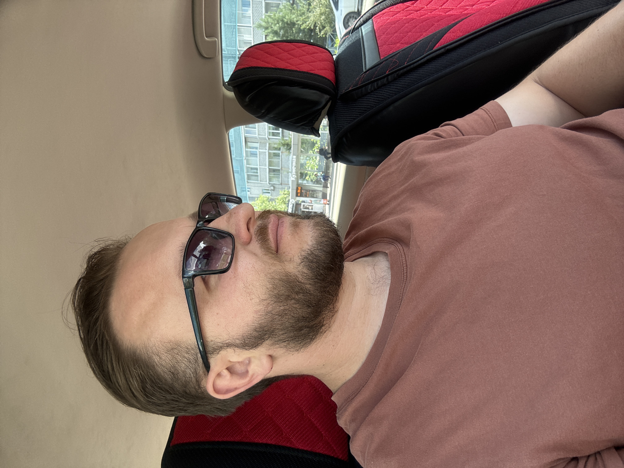 In Blagoveshchensk, Russia, a man with short brown hair, a beard, and sunglasses is sitting in the driver's seat of a car. He is wearing a brown t-shirt. The car has red and black seat covers. Through the car window, a residential building, trees, and a street with pedestrians and a bus stop are visible. The sky is clear and the scene appears to be during daytime.