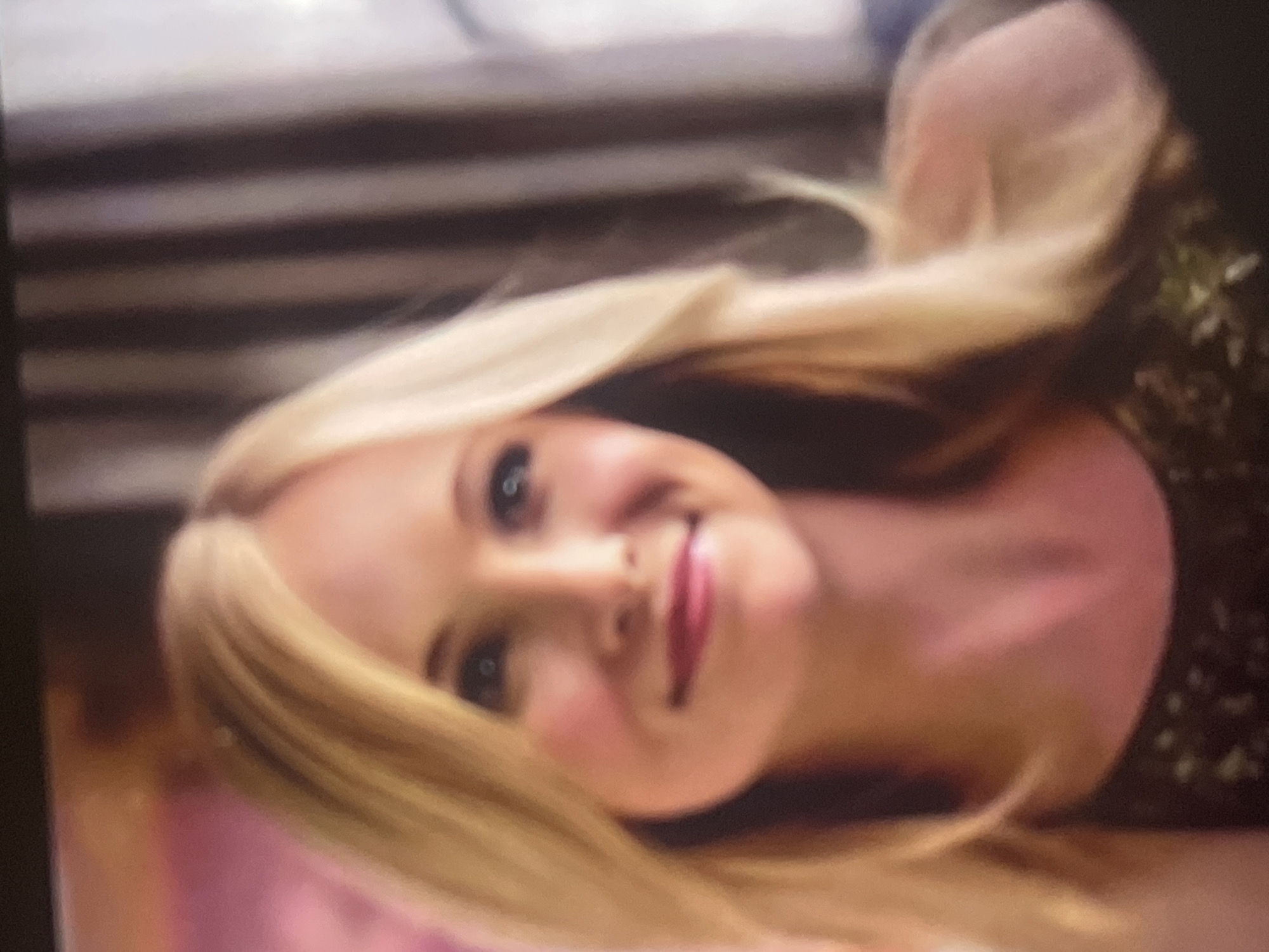 A slightly blurry and rotated portrait captures a young woman with long, straight blonde hair and light blue eyes. She is looking towards the viewer with a gentle, closed-mouth smile, exhibiting a pleasant and content expression. Her cheeks show a light blush, and she is wearing pink or red lipstick. Her hair frames her face and extends over her shoulders. She appears to be wearing a dark top, possibly with a subtle patterned or embellished texture visible on the right side of the image.
The background is out of focus, indicating an indoor setting. To the left, blurred horizontal dark stripes suggest features like window blinds or a wooden wall. A faint pink object or surface is partially visible at the bottom left. The image quality suggests it might be a photograph of a screen. There are no specific location cues visible in the image itself to confirm Poltava, Ukraine, nor is there any visible text. The time of day and weather are not discernible.