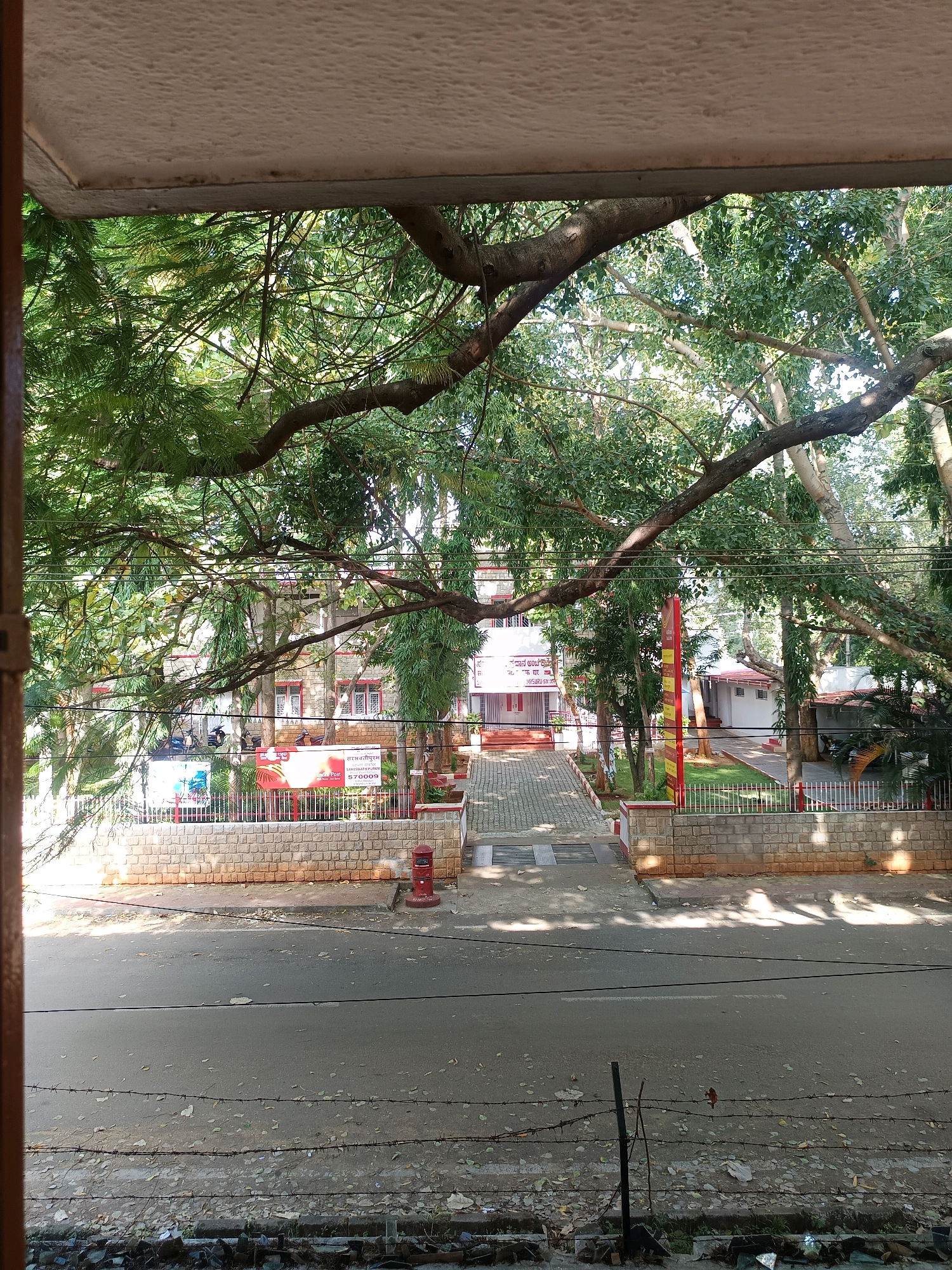 The image depicts a view from an elevated position, looking out towards a building complex with a large tree in the foreground. The tree's branches spread across the frame, casting shadows on the scene below. In the mid-ground, a building with a red-tiled roof and light-colored stone walls is visible. A paved walkway leads up to the entrance of this building, flanked by a low stone wall. Signs with text in a non-English script are posted near the entrance. To the right of the building, there is a smaller white structure and a grassy area. A red postbox is situated on the sidewalk near the road. The foreground consists of a road with some litter and debris, separated by barbed wire fences. The overall environment appears to be a sunny day in an urban or semi-urban setting, possibly in India, given the presence of the postbox and the signage. There are no people visible in the image.