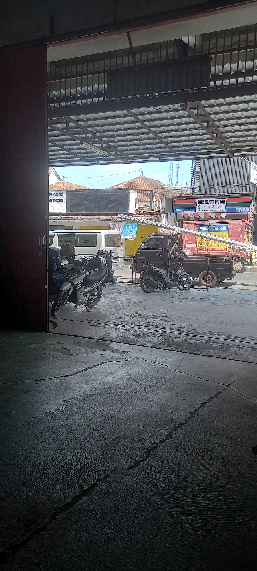 The image depicts an outdoor scene in Banjarnegara, Indonesia, likely a commercial area with a covered workshop or garage entrance in the foreground. The perspective is from inside a building, looking out onto a street. In the foreground, a concrete floor with visible cracks and water stains is visible. To the left, a dark red sliding door is partially open, revealing the interior of the building.
The outdoor scene features several vehicles. Two motorcycles are parked on the left side of the frame, one silver with the "Yamaha" logo visible, and another black motorcycle parked behind it. Further back, a white van is parked. To the right of the van is a yellow vehicle, possibly a truck or van. A black flatbed truck is parked in the center, carrying long metal pipes. On the right side of the truck, a black motorcycle is parked.
Behind the vehicles, there are buildings with tiled roofs. Signs are visible on some of these buildings. One sign, in red and green, reads "SUKSES JAYA MOTOR" and below it, "Bengkel Motor Harga Hemat". Another sign in blue and white advertises "LAUNDRY". Further to the left, a sign with white text on a black background reads "ER GOOD OD SHOES". In the background, several communication towers are visible against a clear blue sky. The overall impression is a busy commercial street in an Indonesian city.