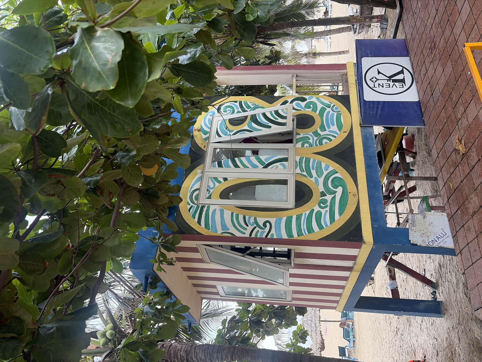 A brightly colored, small building on stilts sits on a sandy beach, partially obscured by lush green foliage from overhead trees. The building's walls feature vertical red and white stripes, and the front is adorned with a large, yellow, green, and blue swirling pattern around two windows. The windows are open, revealing the interior. A blue platform is attached to the front of the building, displaying a white square sign with a black circular logo featuring the letter 'K' and the word 'EVENT'. In the background, the ocean is visible, with beach umbrellas, lounge chairs, and palm trees indicating a tropical location. The scene appears to be in the city of Đà Nẵng, Vietnam, likely a beachfront area.