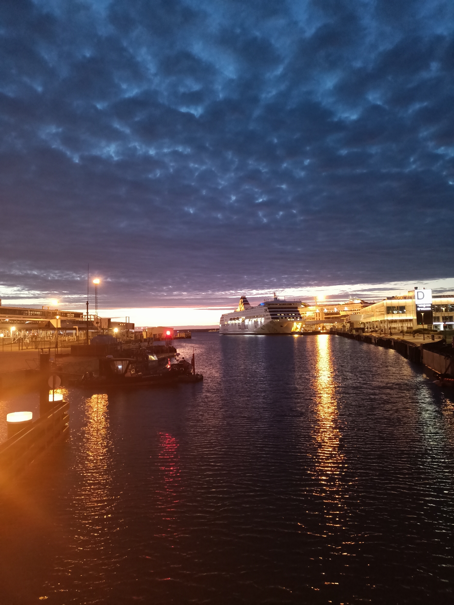 A wide-angle, eye-level, full shot captures the harbor of Tallinn, Estonia, at dusk. The sky is filled with dramatic, dark, rippling clouds, illuminated by hues of deep blue, gray, and streaks of purple and orange near the horizon. A large ferry, labeled "TALINK SILJA LINE," is docked at a terminal on the right side of the frame, its lights reflecting on the water. In the foreground, a smaller boat with red lights is moored near the left bank. The water is calm, with shimmering reflections of the lights from the ferry, terminal buildings, and harbor lights, creating a warm, golden glow. The terminal buildings are modern, with illuminated windows and signage, including a large "D" visible on one structure. The overall scene is serene and evocative, portraying the quiet transition from day to night in a bustling port city.