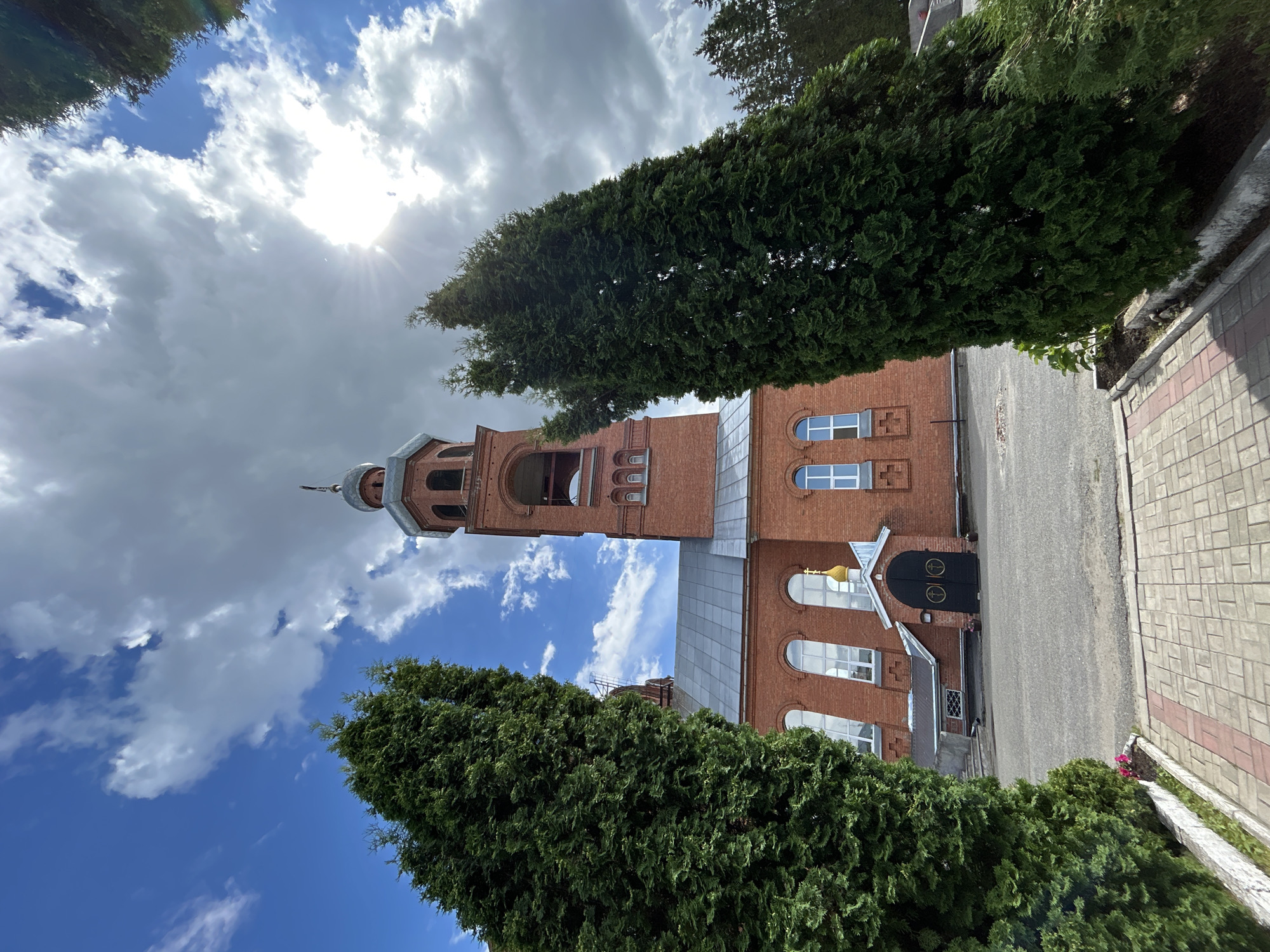 The image shows an exterior view of a brick church with a bell tower in Medvedevo, Russia. The church has a grey roof, arched windows, and a golden dome. A large black double door serves as the entrance. Two tall, dense evergreen trees frame the foreground, with a paved walkway leading towards the church. The sky is partially cloudy with the sun visible and casting rays. The ground in front of the church is asphalt.