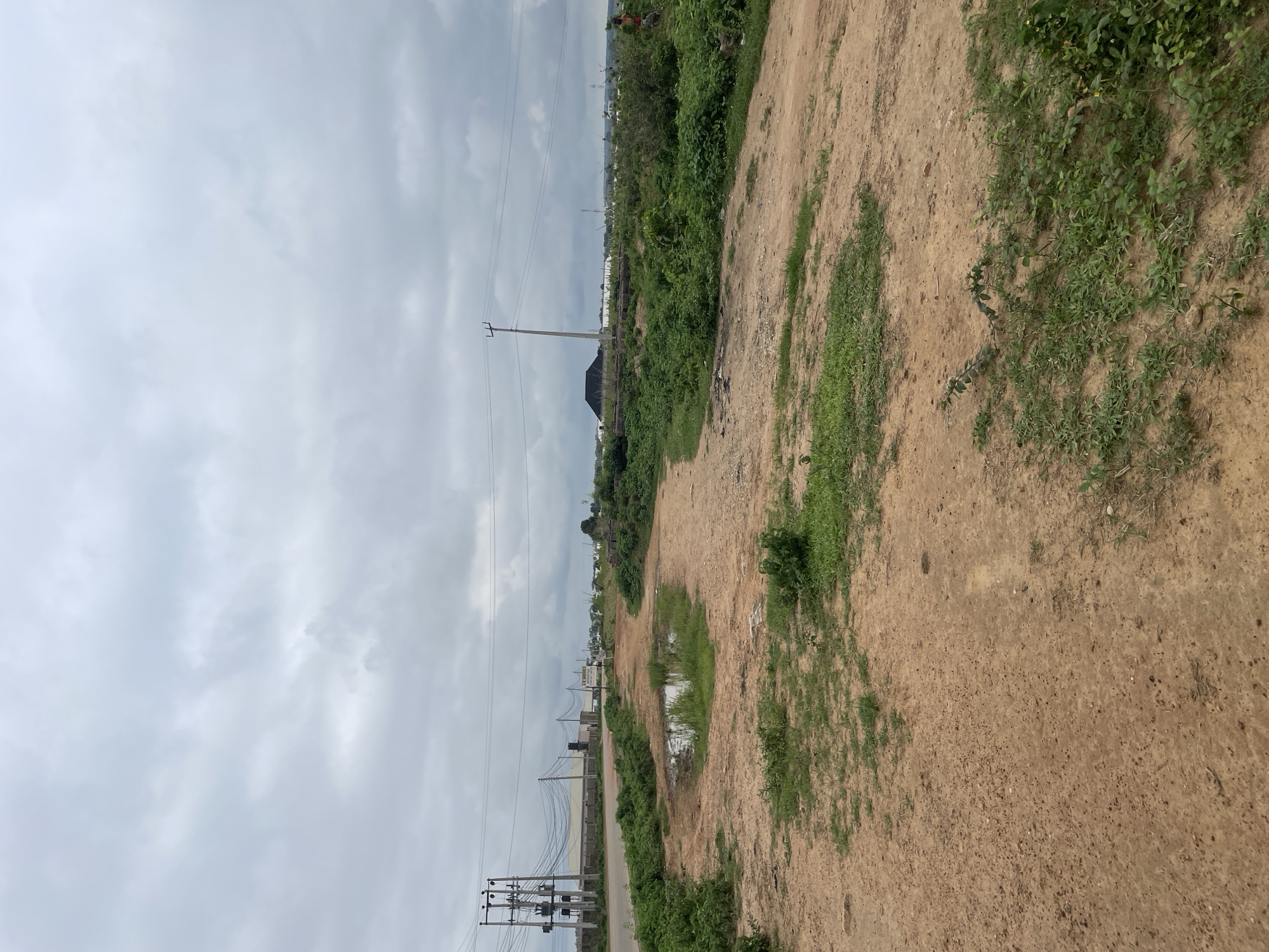 The image shows an overcast sky over a predominantly rural or undeveloped area. In the foreground, there is a dirt path with patches of grass and a small puddle of water. The path leads into a landscape characterized by dense green vegetation, particularly on the left and right sides of the frame.
In the middle ground, a paved road runs horizontally across the scene. Along this road, there are several power poles with transformers and numerous electrical wires stretching across the frame. In the distance, there are various structures, including what appears to be a large industrial building or warehouse. Further in the background, there are more scattered buildings and some distant hills or mounds of dark material, possibly coal or earth. A utility pole stands in the center of the frame, supporting power lines.
On the far right side of the image, a person wearing a red shirt is visible, standing amidst the vegetation. The overall impression is one of a developing area where infrastructure like roads and electricity are present, but the land is still largely undeveloped with natural vegetation. The location is identified as Baganakwo, Nigeria.