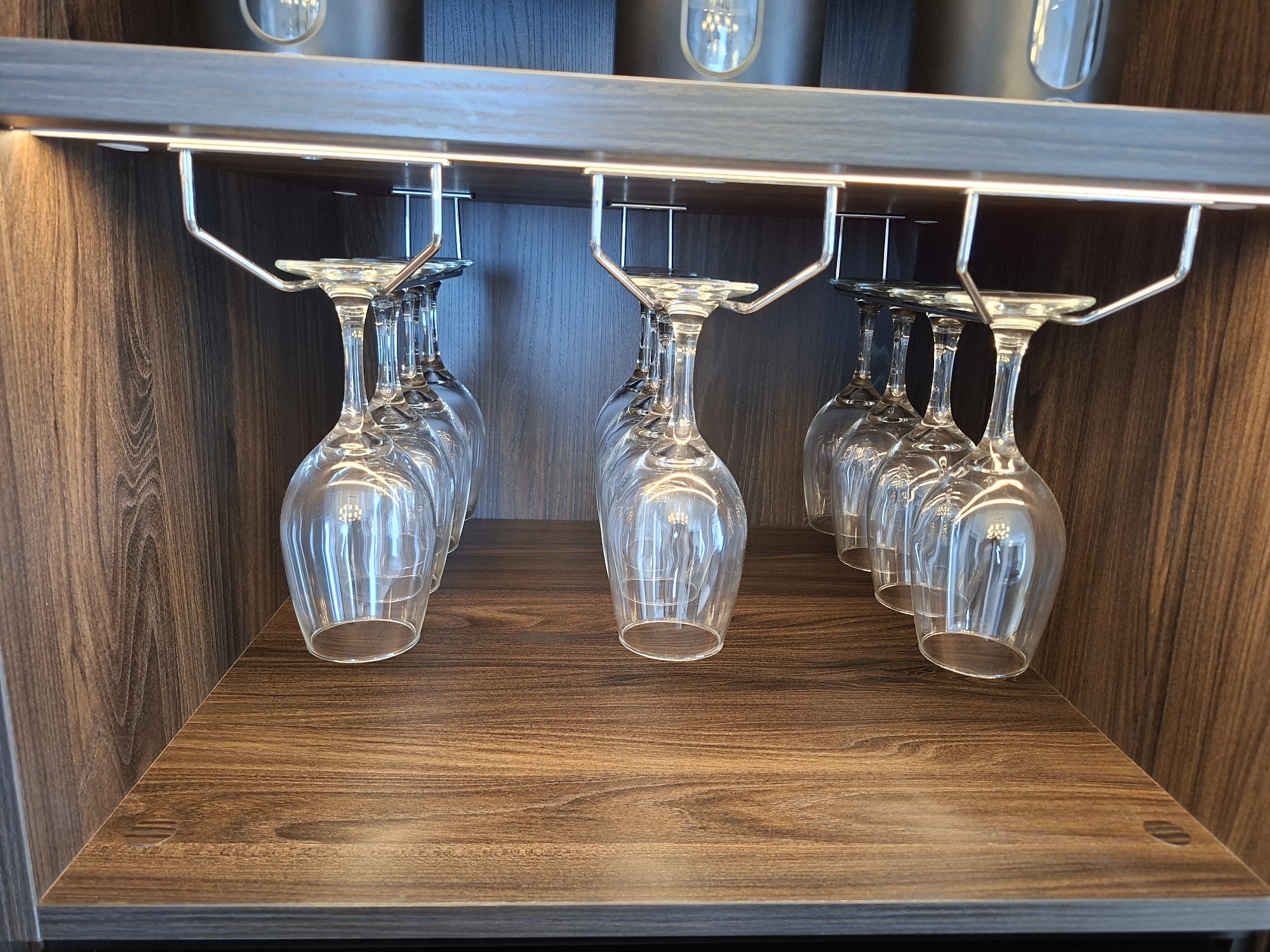 A dimly lit, modern wine rack features several clear wine glasses hanging upside down from a metallic rack. The rack is mounted beneath a shelf that has integrated LED lighting, casting a warm glow. The background consists of dark wood-paneled walls, creating a sophisticated and contemporary ambiance. The wine rack is positioned on a wooden shelf with a visible wood grain pattern. The image is taken from a slightly elevated angle, focusing on the arrangement of the wine glasses. The scene is devoid of any people or specific indicators of its location in West Jakarta, Indonesia.
