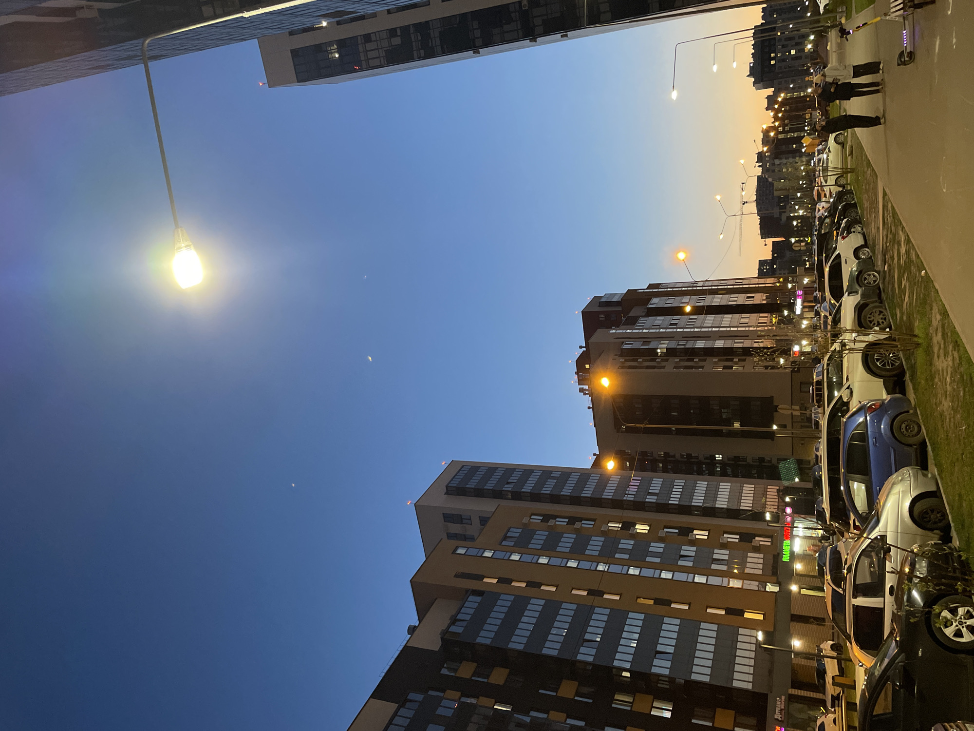 The image captures a street scene in Kazan, Russia, at dusk. Tall residential buildings line both sides of the street. A row of parked cars fills the right side of the frame, facing towards the top of the image. Several people are standing on the sidewalk, engaged in conversation. Streetlights illuminate the scene, casting a warm glow on the buildings and cars. The sky is a gradient of blue and orange, indicating the time of day.