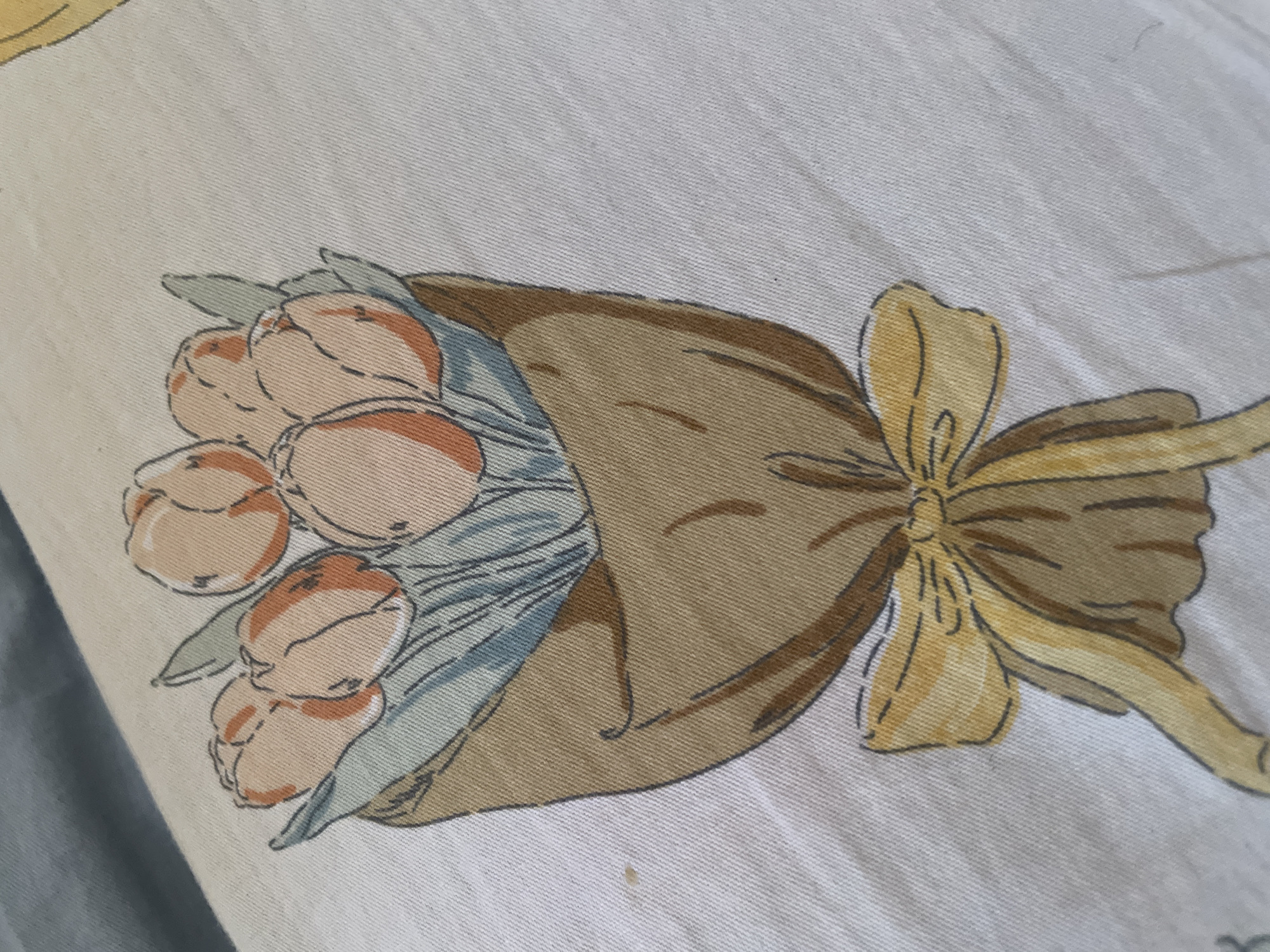 The image shows a close-up view of a fabric with a printed illustration of a bouquet of tulips. The tulips are depicted in shades of pale orange and pink, with green leaves. The bouquet is wrapped in brown paper, tied with a yellow ribbon. The fabric appears to be a light color, possibly white or off-white, and has a textured weave. The image is taken from a slightly elevated angle, and the lighting suggests an indoor setting. There is no visible indication of a specific location within Tashkent, Uzbekistan, or any people or actions.